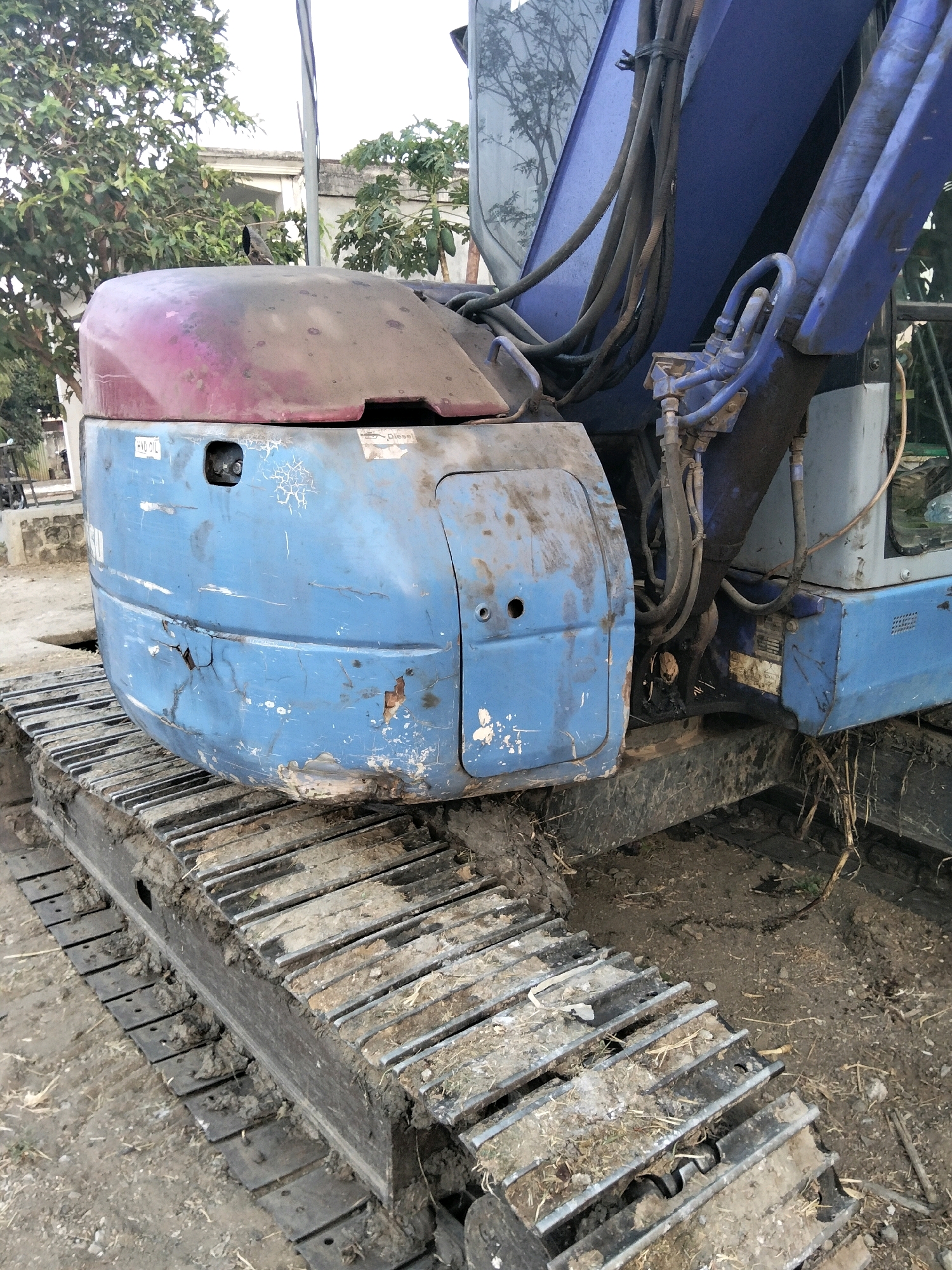 The image shows a close-up, slightly angled view of the front portion of a blue excavator. The excavator is situated outdoors on a dirt surface, with some dry vegetation and debris present. The tracks of the excavator are visible, covered in dirt and mud. The front panel of the excavator is blue with significant signs of wear and tear, including cracks, scratches, and patches of rust. A pinkish-red colored cover is visible on top of the blue panel. There are several stickers on the blue panel, including one with "HYD OIL" and another with "Diesel". A dark-colored bird is perched on the pinkish-red cover. In the background, trees, foliage, and parts of a building are visible, suggesting an urban or semi-urban environment in Mrican, Indonesia. The overall impression is that of a weathered and used piece of heavy machinery.