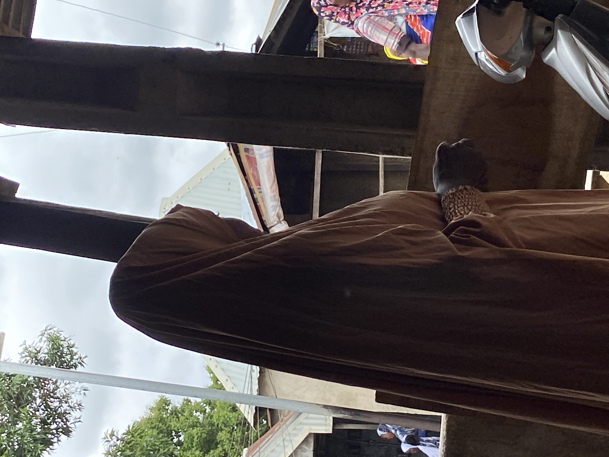 The image appears to be taken in an outdoor market or commercial area in Mararaba, Nigeria. The scene is partly obscured by a beam or part of a building structure in the foreground. A person covered in a brown cloth is visible, with a hand adorned with woven or beaded bracelets resting on the cloth. Adjacent is a motorbike, reflecting sunlight from its headlights. Above and slightly behind the covered person, a woman wearing a colorful patterned top is seen, potentially engaging in commerce. A corrugated metal roof suggests a market stall or building. Further back, there are trees and structures suggesting a built environment. The sky is overcast.