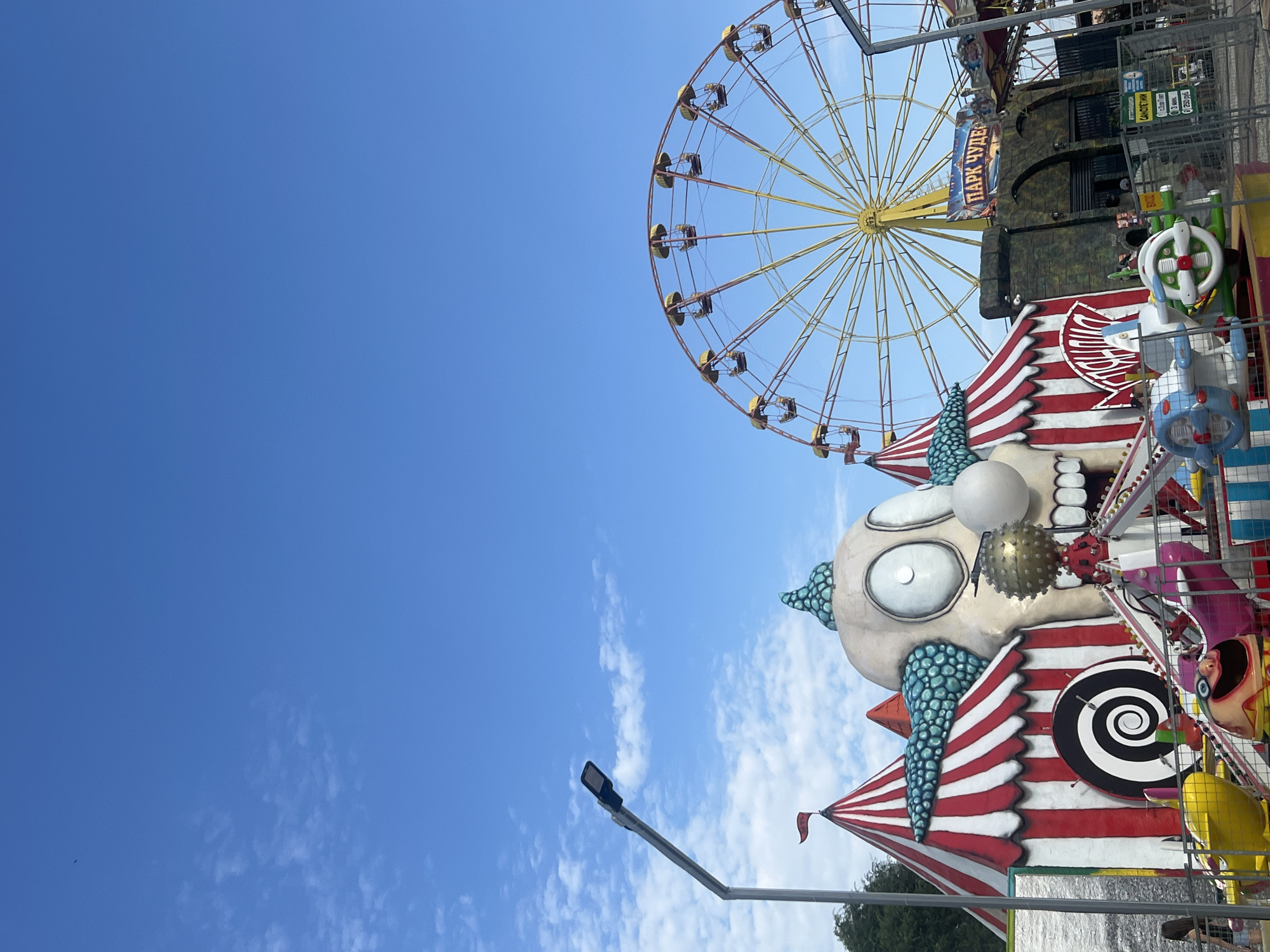 This is a vertical, medium-shot, outdoor photograph taken on a clear day with a blue sky and scattered white clouds. The scene depicts an amusement park in Golubitskaya, Russia, dominated by a large, stylized clown face structure with a gaping mouth, wide eyes, and a bumpy, teal-colored headpiece. The clown is part of a carnival-like setup with red and white striped tents and a ride featuring small, colorful vehicles. In the background, a large Ferris wheel with yellow gondolas is visible, along with a sign that reads "ПАРК ЧУДЕС" (Park of Wonders). A street lamp is present on the left side of the frame. The overall impression is of a vibrant and somewhat unsettling amusement park attraction.