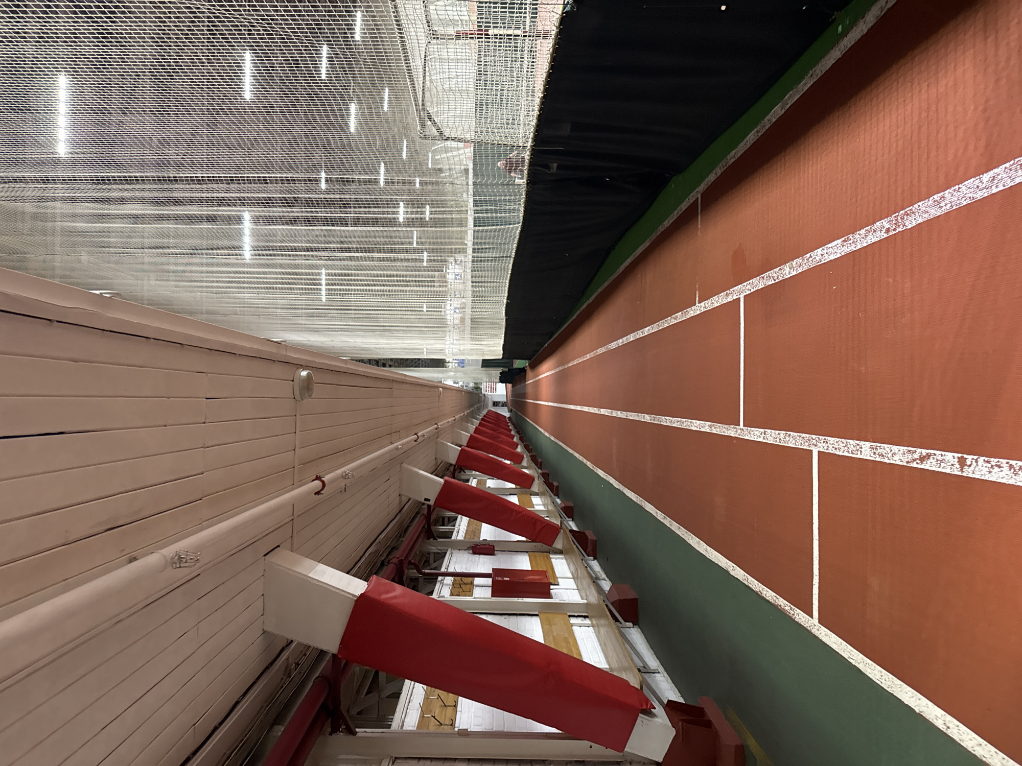 The image depicts an indoor track and field facility. The perspective is from a high vantage point, possibly the upper bleachers. The track itself is a reddish-brown surface with white lane markings. A green border separates the track from a wall on one side. Protective padding with red covers is along the wall. Above the track is a dark, possibly sound-dampening material, and further up is a white netting structure. On the opposite side of the track is a light-colored wall with horizontal paneling. The image does not contain any people, subjects, actions, or interactions. No location in the city of Saint Petersburg, Russia can be determined from the image.