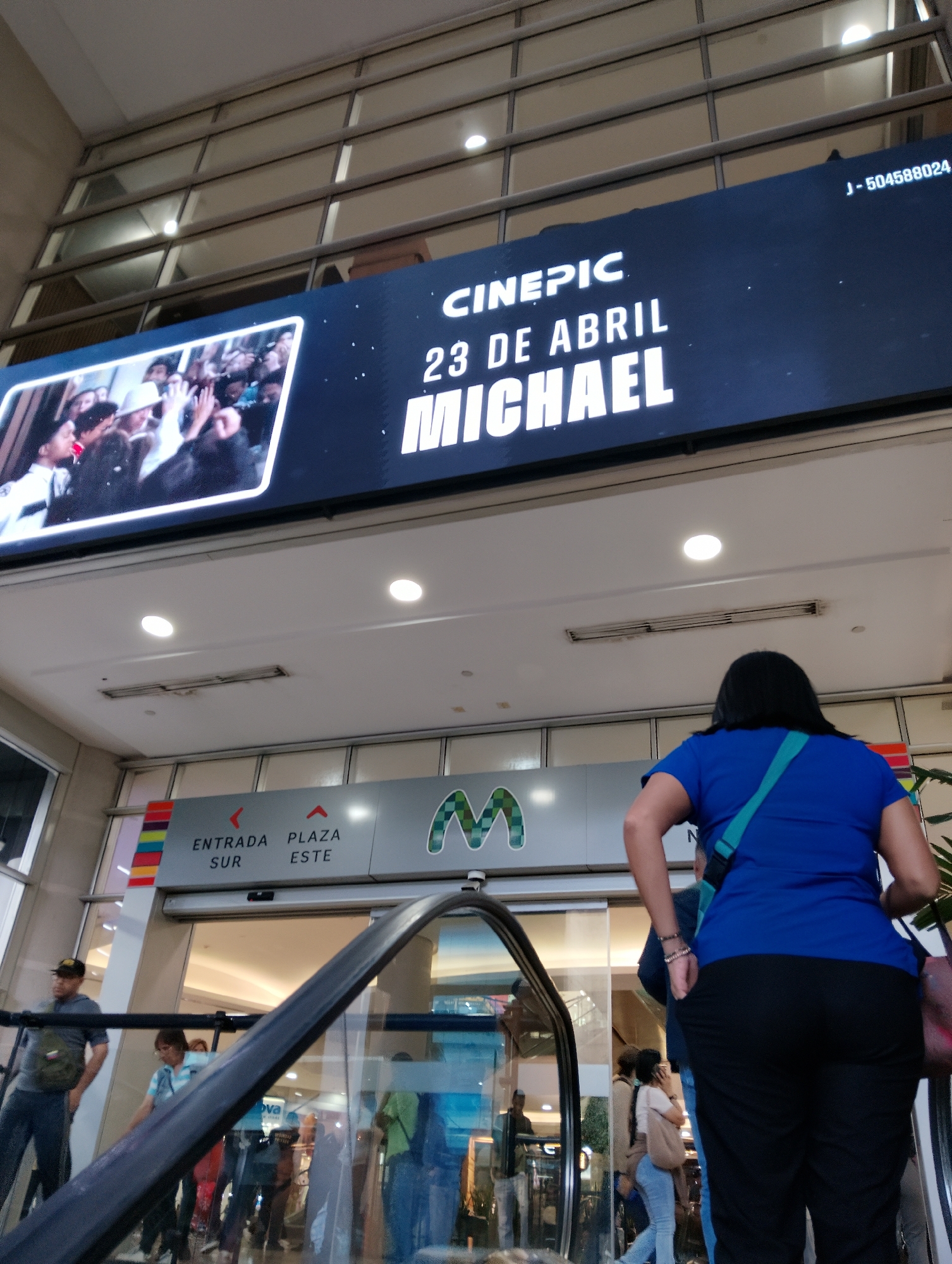 The image shows the interior of a shopping mall in Unknown, Unknown. Above an escalator, a large digital banner displays the word "CINEPIC" followed by "23 DE ABRIL" and "MICHAEL" in bold white letters. To the left of the text, there is a photograph on the banner with multiple people raising their hands. Beneath the banner, there is an entrance to the mall indicated by signage that reads "ENTRADA SUR" and "PLAZA ESTE" flanking a green "M" logo. People are visible both on the escalator and walking through the entrance, including a woman in a blue shirt and black pants who is facing away from the camera. The mall architecture features a wall of window panels.