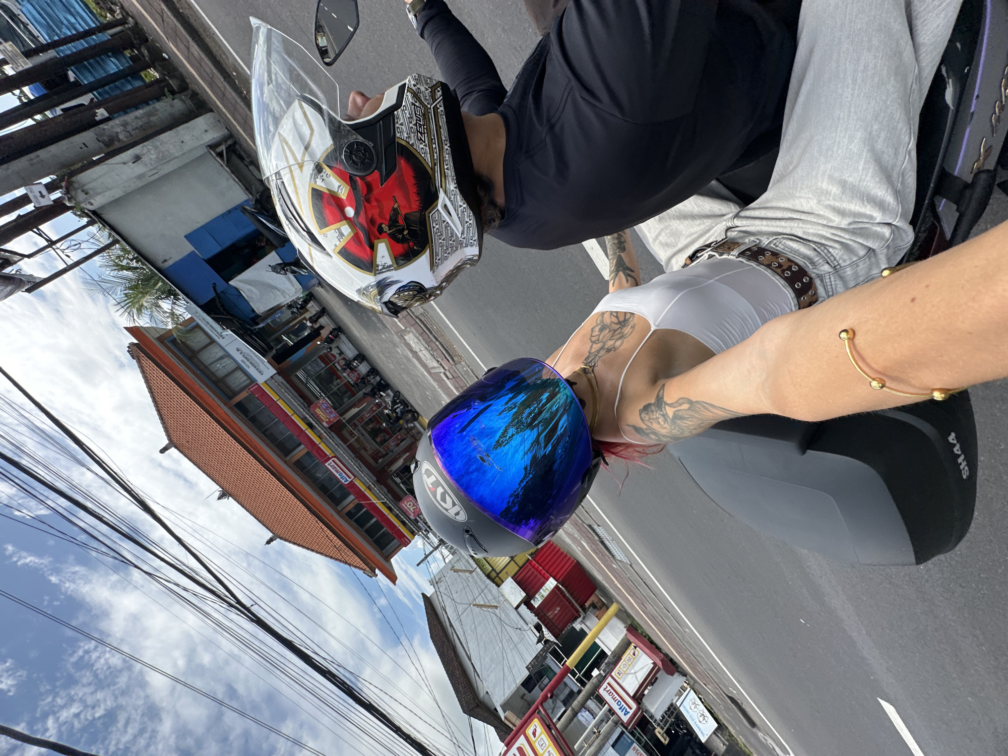 A medium-angle, tilted, outdoor shot captures two people on a parked motorcycle in Kuta Selatan, Indonesia. The perspective is from behind the riders, looking slightly downward at a tilted angle.
The person in the front wears a white and gold motorcycle helmet with a red and black graphic. They are dressed in a black long-sleeved shirt and light gray denim jeans with a brown belt. The passenger in the back wears a gray motorcycle helmet with a blue reflective visor. They are wearing a white top. Tattoos are visible on the arms and upper back of both individuals. A gold bracelet adorns the passenger's left arm. The motorcycle has a black and gray storage case mounted behind the passenger seat.
The background includes buildings and power lines set against a partly cloudy sky. Storefronts with signage in Indonesian are visible. The architecture features red tile roofs and building facades characteristic of the region.