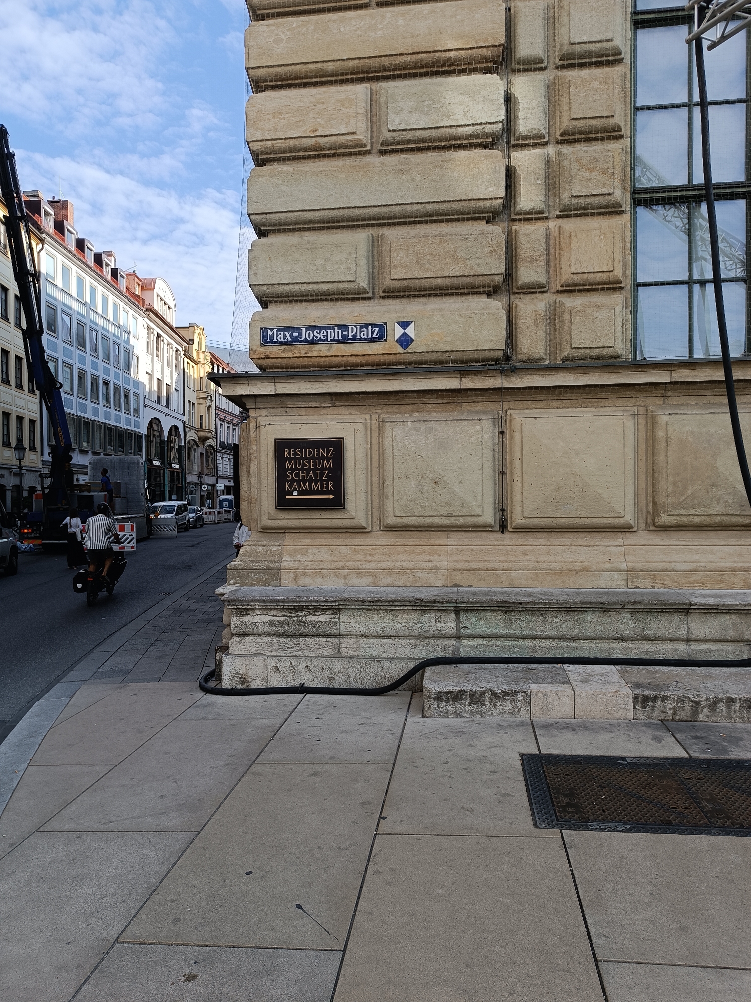 The image shows a street scene in Munich, Germany, with a focus on the corner of a large stone building. A street sign reads "Max-Joseph-Platz". Below this sign, a bronze plaque indicates "RESIDENZ MUSEUM SCHATZKAMMER" with an arrow pointing to the right. The building's facade is made of large, carved stone blocks, and a fine mesh netting covers parts of it. A window with a grid pattern is visible on the right side of the building.
In the foreground, a sidewalk made of large concrete slabs is visible, with a dark, thick cable running along the base of the building and across the sidewalk. A metal manhole cover is set into the sidewalk.
On the street, a person is riding a scooter, wearing a striped shirt and shorts. A large crane arm extends from the left side of the image, obscuring some of the background buildings which appear to be in a European architectural style. Other vehicles and pedestrians are visible further down the street, which appears to be an urban commercial area. The sky above is blue with scattered clouds.