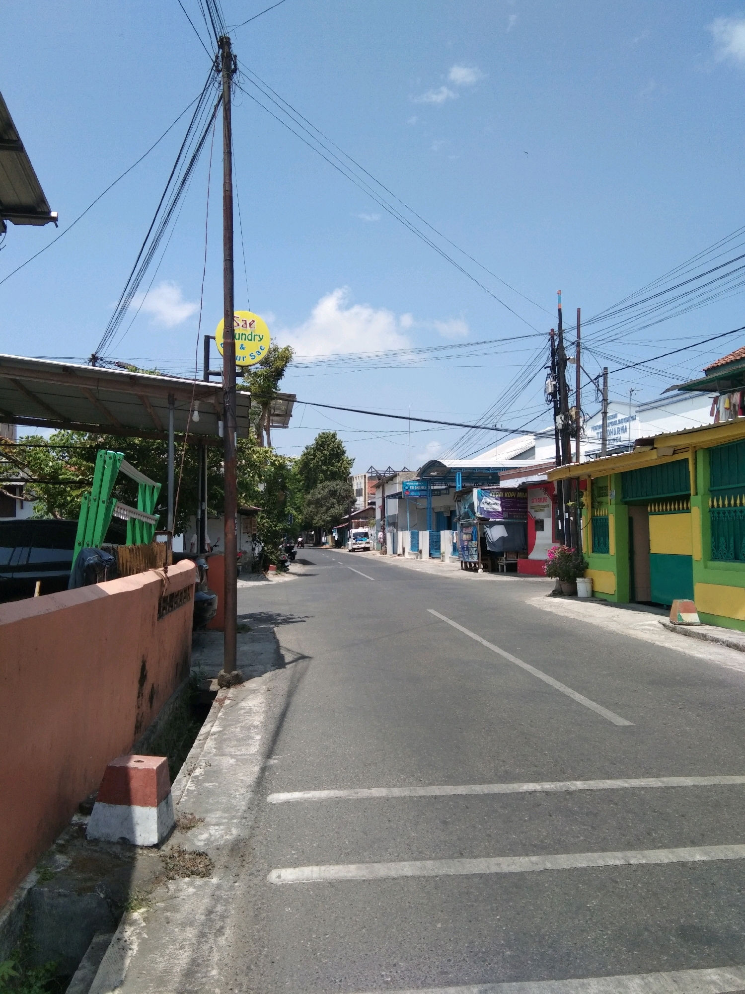 This is a low-angle, eye-level, full shot of a street in Kebumen, Indonesia, under a clear blue sky with scattered clouds. The perspective is from the side of the road, looking down the street. The street is paved with asphalt and has white lane markings. On the left side of the street, there is a terracotta-colored wall with a concrete sidewalk and a drainage ditch. A red and white concrete marker is placed at the edge of the ditch. Above the wall is a covered structure with a green ladder leaning against it. A utility pole with many tangled wires stands prominently on the left. A yellow circular sign with Indonesian text, possibly advertising a laundry service, is attached to the pole. Further down the street, there are various buildings with colorful facades, including green and yellow. Some of these buildings appear to be shops or businesses, with signs visible. A white delivery truck is parked on the right side of the street in the distance. The overall scene depicts a typical urban street in Indonesia on a sunny day.