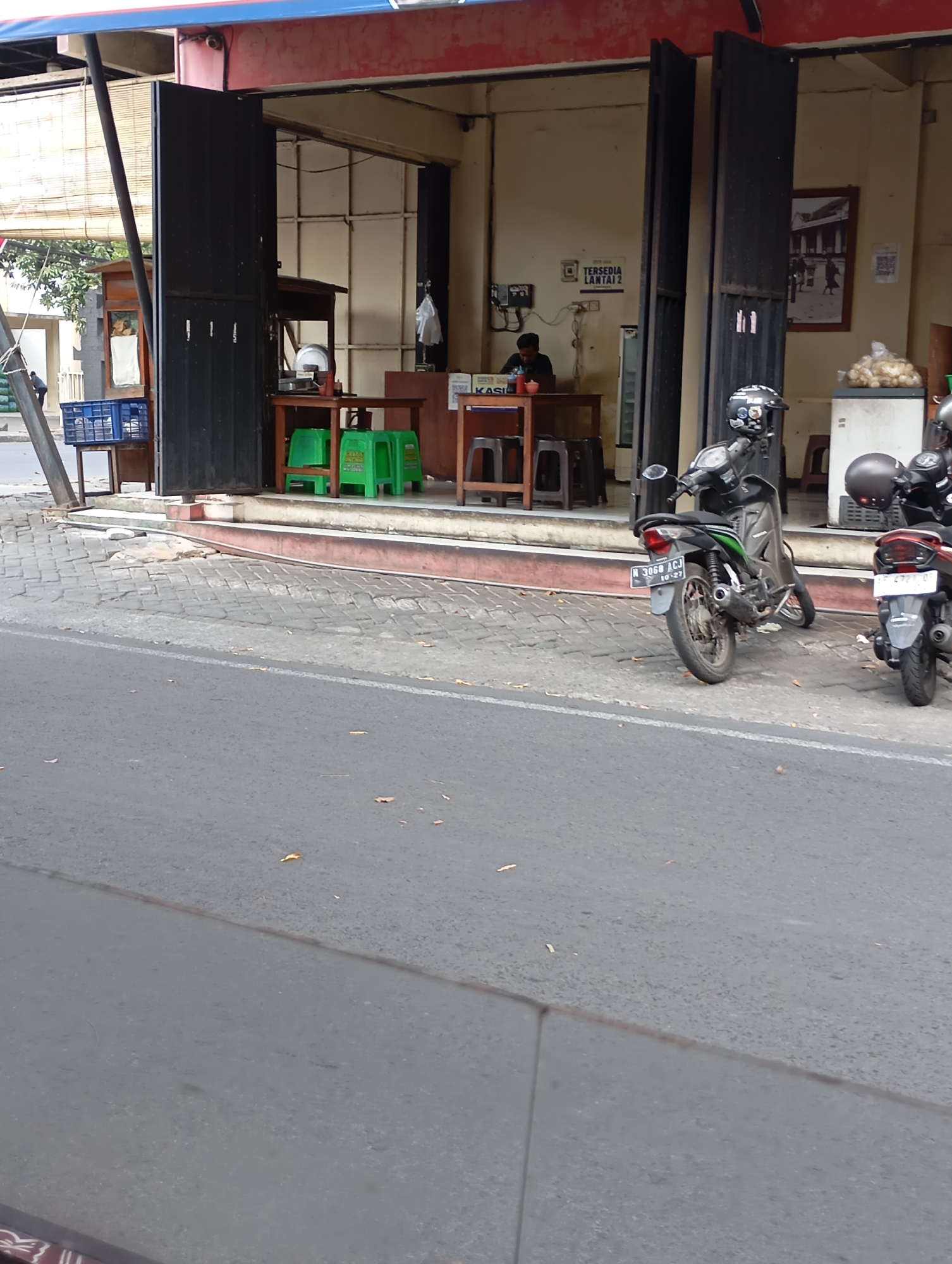 The image depicts a street scene in front of a shop in Malang, Indonesia. A man is seated at a table inside the shop, which has an open storefront with several green chairs and tables visible. Two motorcycles are parked on the sidewalk outside the shop. The street in the foreground is paved with asphalt. The shop appears to be a food establishment, with a sign indicating "Tersedia Lantai 2" which translates to "Available Second Floor".