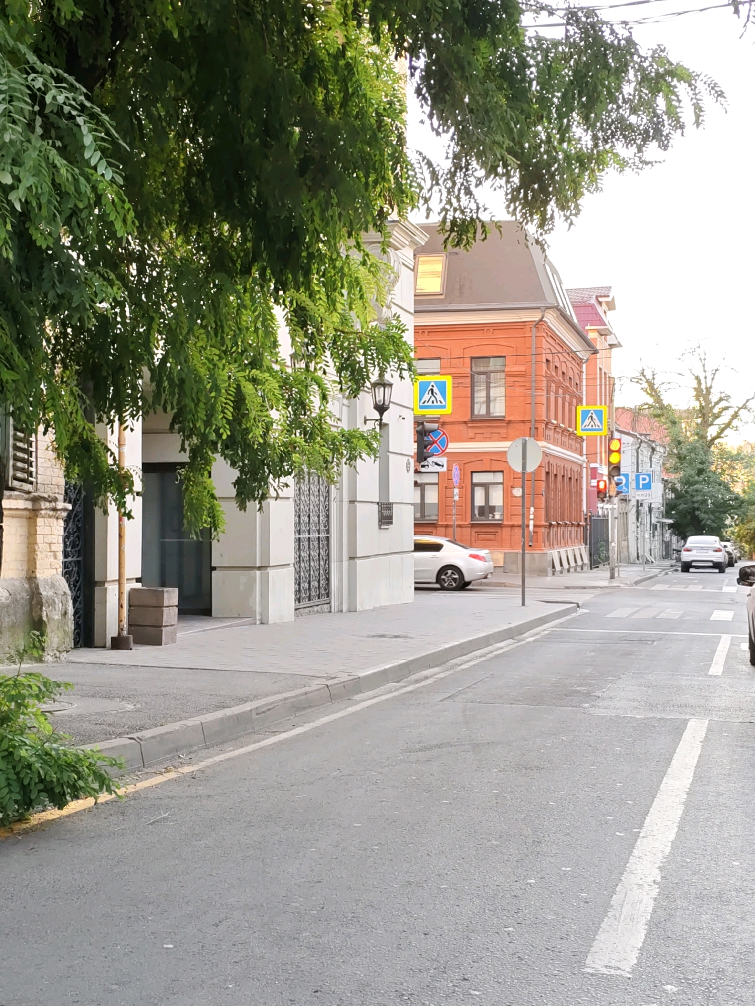 A street scene in Rostov-on-Don, Russia, features a pedestrian crossing with two yellow pedestrian crossing signs. On the right side of the street, a traffic light shows a red signal. A white car is parked on the right side of the street, and another white car is visible crossing the street in the distance. Buildings line the street on the left and right, with a red brick building prominently featured on the right. Green trees with lush foliage hang over the sidewalk on the left side of the image. The sky is overcast.