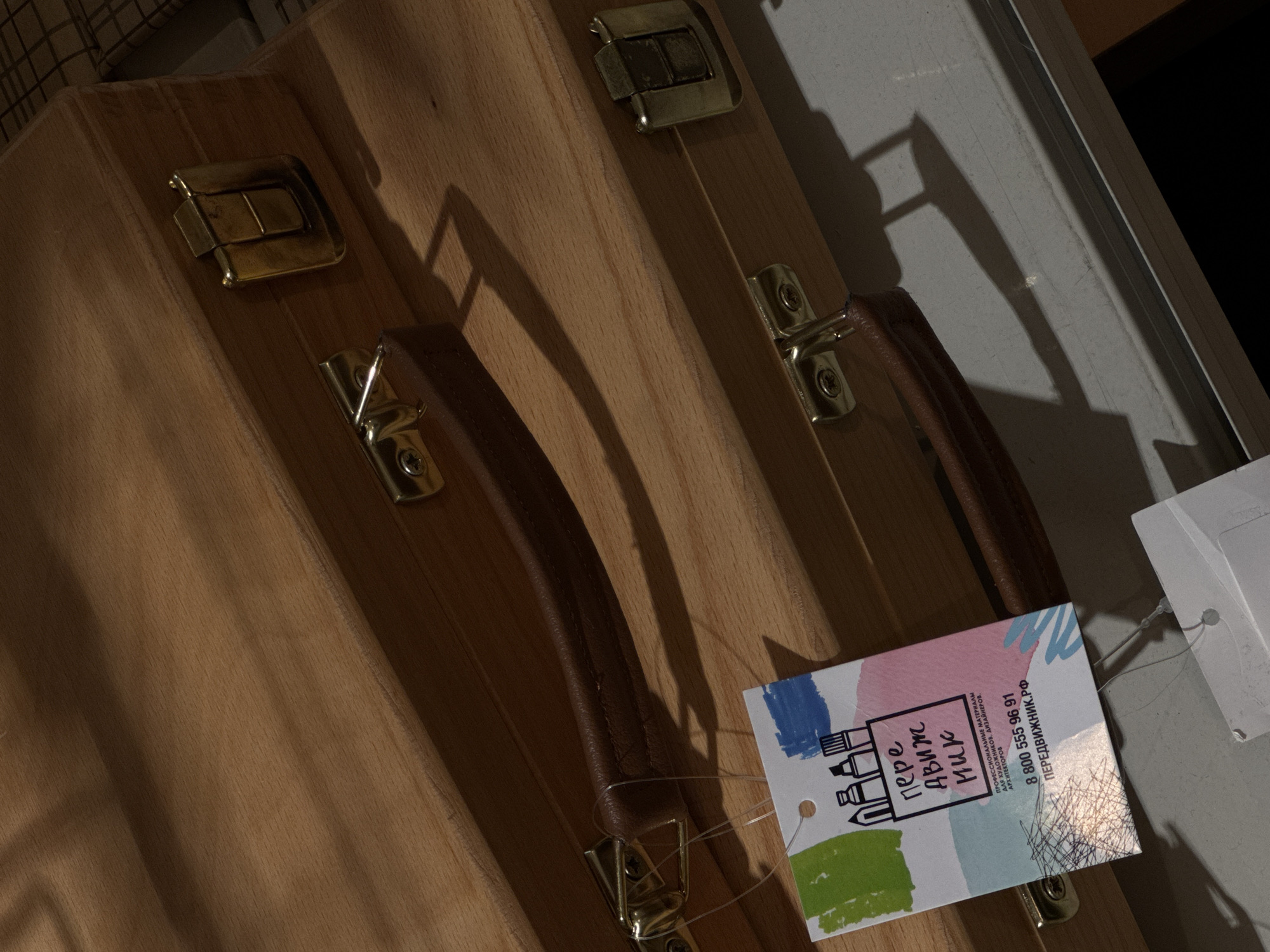 This is a close-up, eye-level, medium shot of two stacked wooden boxes with leather handles and brass latches. The boxes are positioned at an angle to the viewer, with the top box casting a shadow on the bottom box. A white tag with colorful abstract shapes and text in Russian is attached to the bottom box by a clear plastic string. The text on the tag reads "передвижник", "ПРОФЕССИОНАЛЬНЫЕ МАТЕРИАЛЫ ДЛЯ ХУДОЖНИКОВ, ДИЗАЙНЕРОВ, АРХИТЕКТОРОВ", "8 800 555 96 91", and "ПЕРЕДВИЖНИК.РФ". The background is blurred and appears to be a light-colored surface with a shadow. The lighting is dim, creating strong contrasts and shadows. No people or discernible Moscow landmarks are visible.
