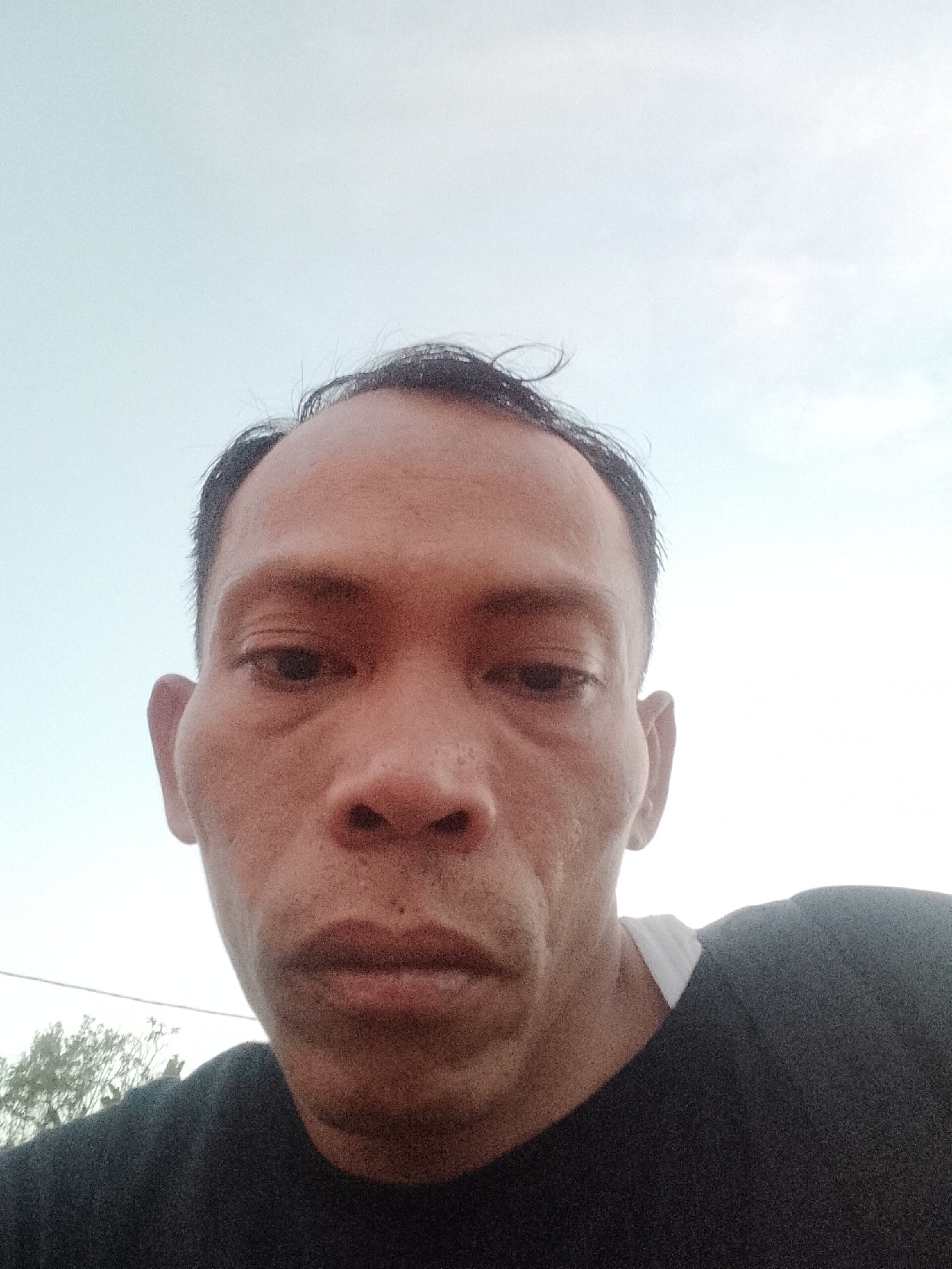 A close-up, low-angle, outdoor selfie taken in Muara Sungsang, Indonesia, shows the upper torso and face of an adult male. The man has dark hair, dark eyes, and medium-toned skin. He is wearing a dark t-shirt. The background is a pale, hazy sky with some indistinct tree foliage visible in the lower left corner. The image is somewhat grainy and has a bright, washed-out quality, possibly due to strong sunlight. The man's expression is neutral, and he is looking directly at the camera. The focus is on his face, with his ears and the top of his shoulders also visible.