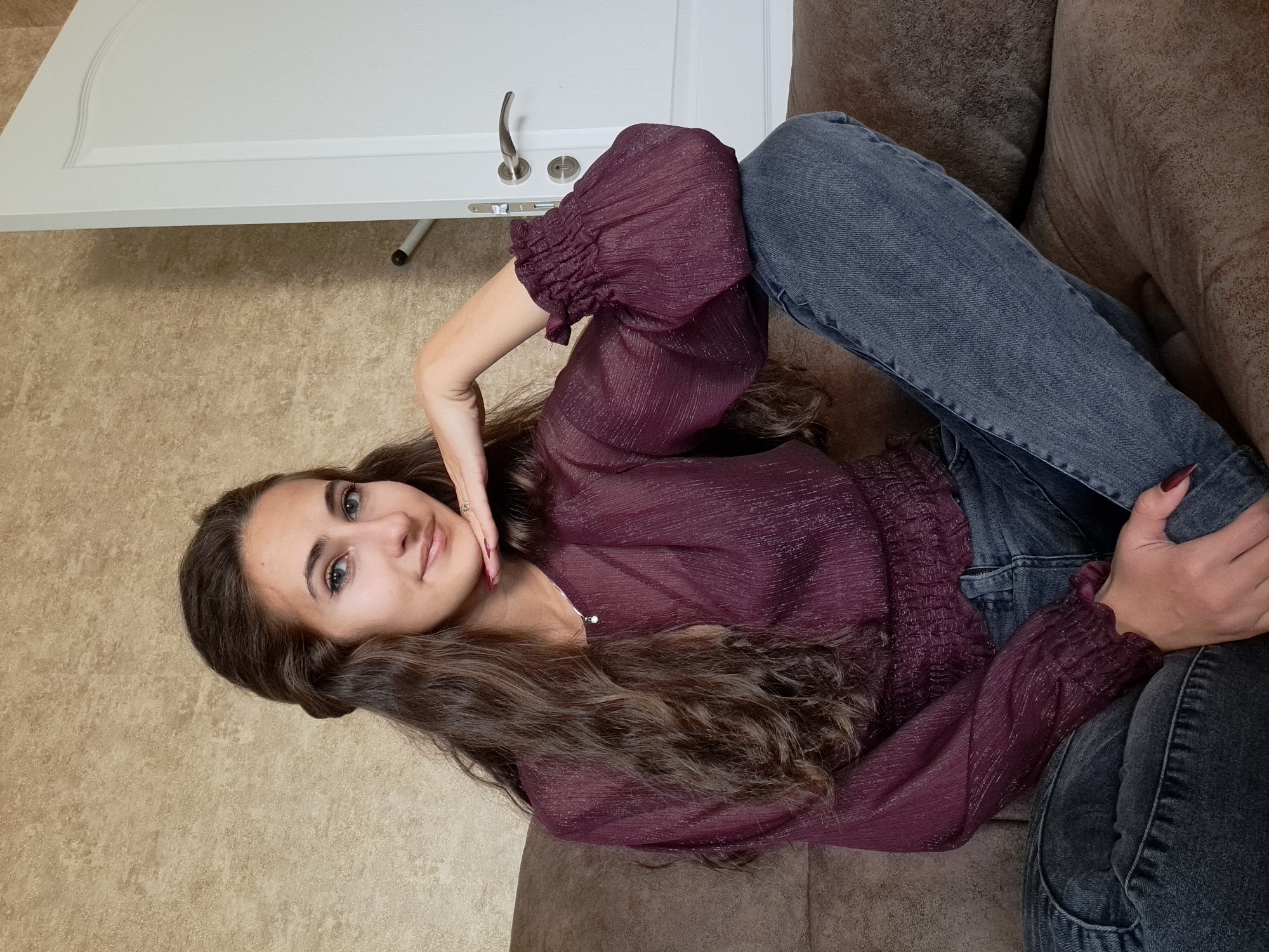 A young woman with long, wavy brown hair is reclining on a brown sofa. She is wearing a dark purple, long-sleeved, textured blouse and gray jeans. Her legs are bent, and one hand rests on her knee while the other is near her face, touching her chin. A white door with silver hardware is visible in the background, along with a beige wall. The location is not evident. No information suggests it is Negovan, Bulgaria.