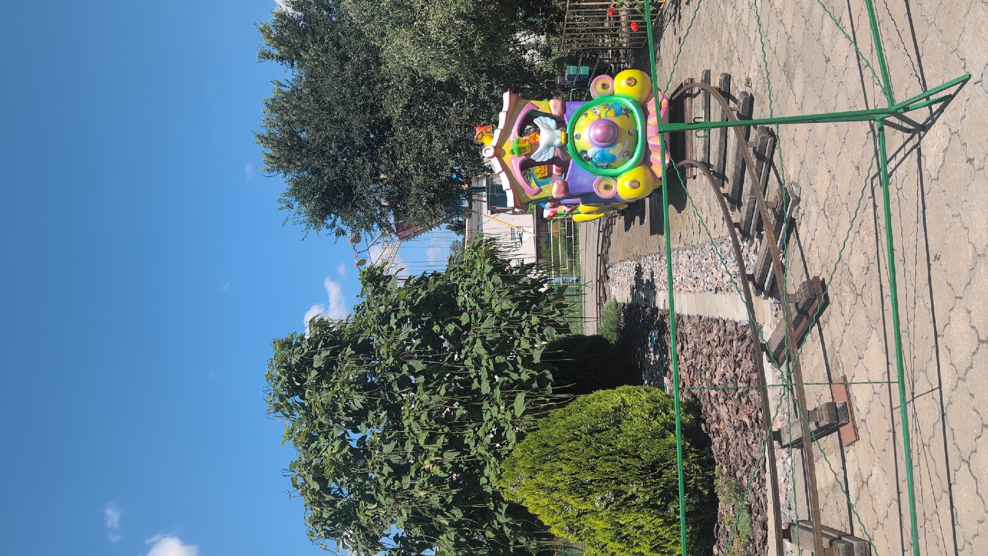 The image shows a children's amusement park ride, a colorful toy train, with a Ferris wheel in the background. The train is yellow, purple, and pink, and features cartoon characters on its front. It is on a small set of tracks in what appears to be a public park. The scene is set on a sunny day with a clear blue sky. The location is in the city of Izmail, Ukraine.