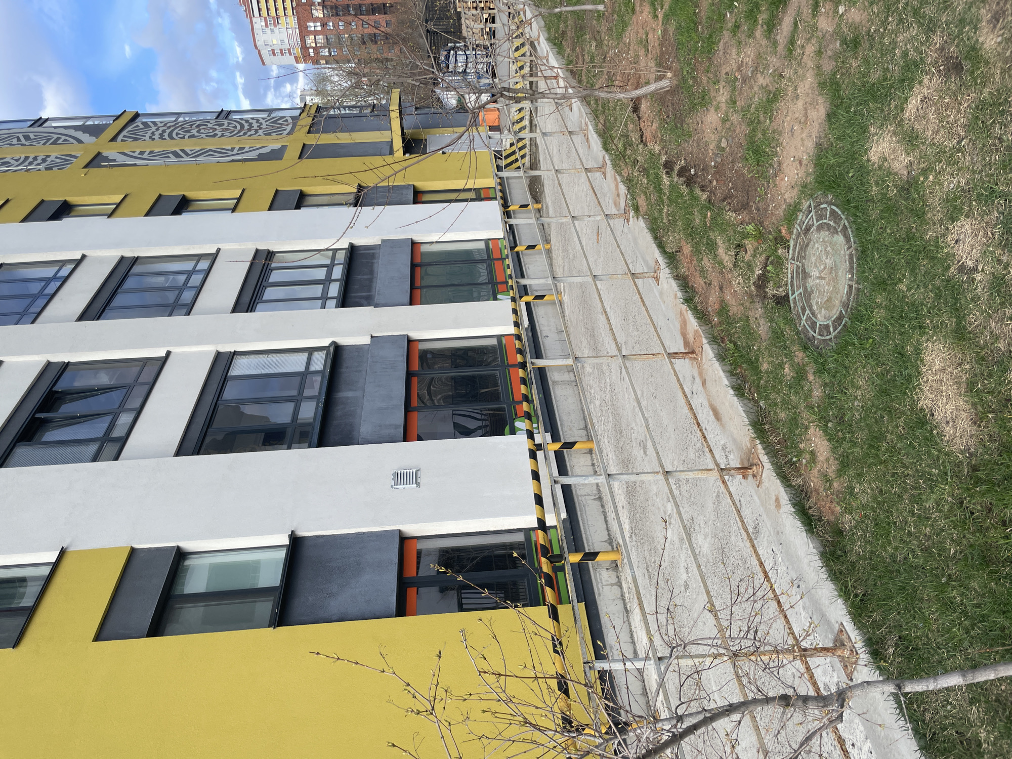 A modern, multi-story building with a vibrant, multi-colored facade dominates the scene. The lower and uppermost sections of the building are a bright yellow, while the middle section is white. Dark grey or black panels frame the numerous rectangular windows and divide sections of the facade. Some window recesses or balcony areas feature bright orange accents. The top yellow section of the building is adorned with large, light-colored geometric and circular patterns.
In the foreground, a concrete sidewalk or path runs parallel to the building. Along the edge of this path, a temporary barrier system composed of metal poles wrapped with yellow and black striped hazard tape is visible. Beyond the path, there is a strip of natural ground with a mix of green grass and patches of bare brown soil. A circular manhole cover with a faint impression of a number or symbol is embedded in the grass. Several bare-branched trees with nascent buds are scattered throughout the foreground and midground, suggesting early spring.
The sky above is partly cloudy blue, indicating daytime with good natural lighting. In the distance, behind the main building, another multi-story building constructed of brick is partially visible. The overall scene depicts an urban environment, likely a recently constructed or renovated area in Izhevsk, Russia. No clear, legible text is visible on the building or other elements.