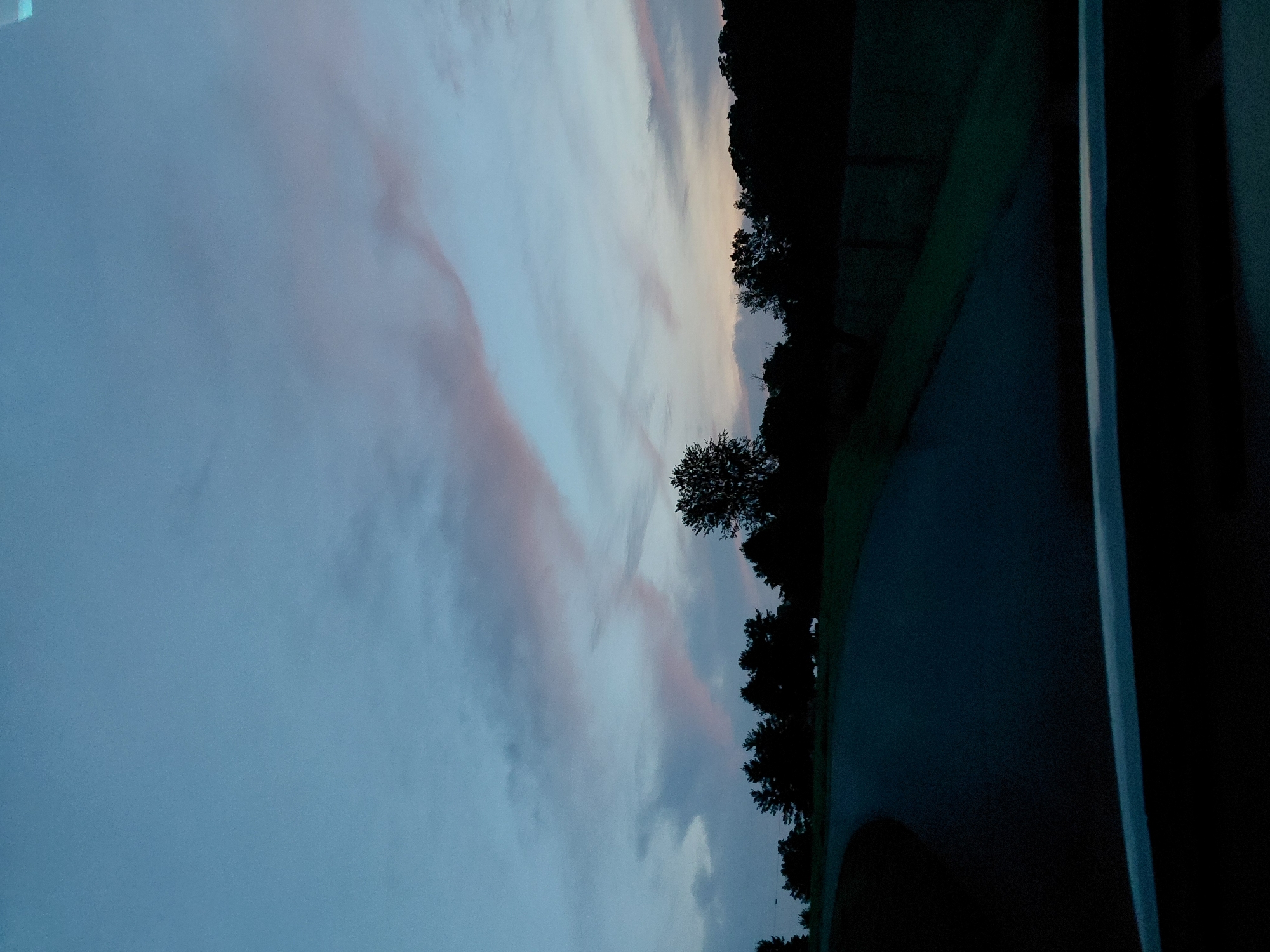 The image shows a road curving through a silhouette of trees against a twilight sky. The sky is a gradient of blue, with wispy clouds tinged with pink. The road appears to be paved and is dark, suggesting it is nighttime or dusk. The trees are densely packed and form a dark line across the horizon. The foreground of the image is dominated by the dark surface of the road and a dark, indistinct object, possibly the dashboard of a vehicle, suggesting the image was taken from inside a car. There are no people visible in the image. The environment appears to be rural or suburban, with no discernible city structures.