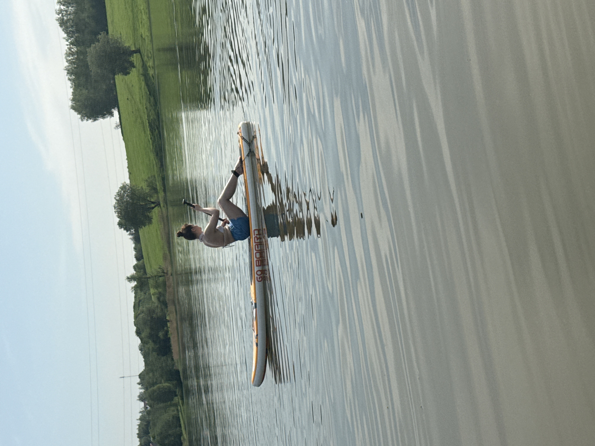 The image shows a person balancing on a paddleboard in a body of water, likely a lake or wide river, in Novosinkovo, Russia. The individual, who appears to be female, is in a partially upright position, with one leg extended. The paddleboard is white and yellow, and the words "GO BOARD" are visible on its surface. The water's surface is rippled, reflecting the surrounding trees and sky. The background includes green fields and trees along the shoreline. A power line can be seen on the left side of the image.