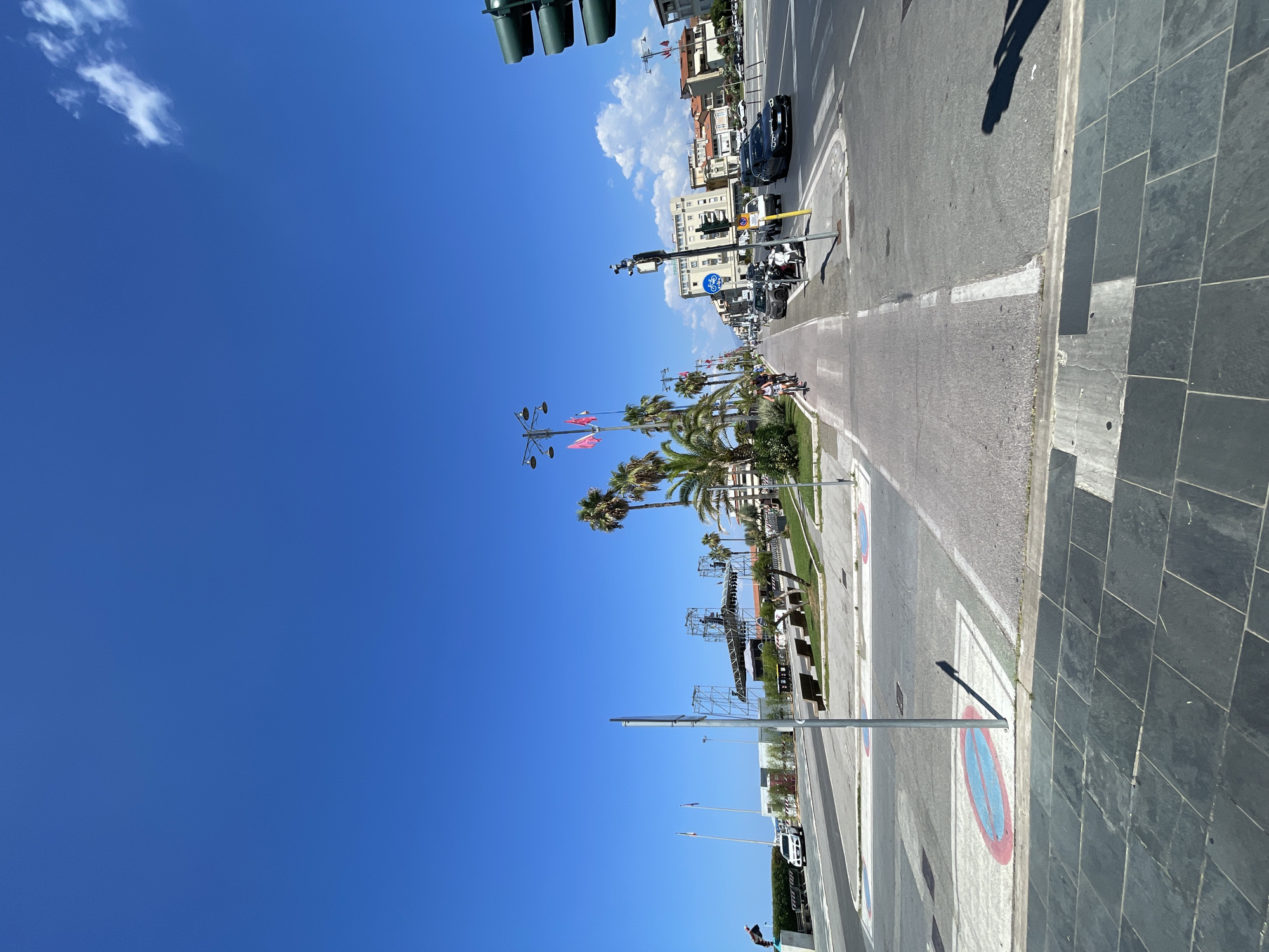 The image shows a wide, sunny street scene in Viareggio, Italy, likely along the waterfront promenade. The sky is a clear, vibrant blue with a few wispy clouds. On the left side of the road, a dedicated bike lane runs parallel to the street, with palm trees and benches lining the walkway. A few people are seen cycling in the bike lane. Across the street, there are buildings, palm trees, and more vehicles. A large stage structure is visible in the background on the left, suggesting a possible event or performance. Traffic lights and street lamps are also present. The overall impression is of a lively, coastal urban environment on a bright summer day.