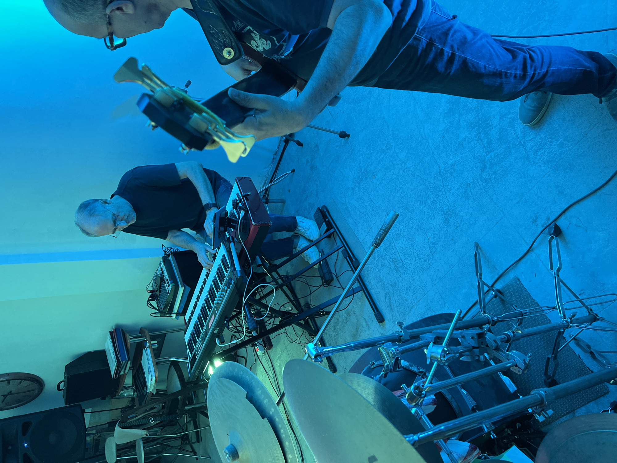 This image captures an indoor musical performance or rehearsal space located in Santeramo in Colle, Italy. The scene is illuminated by a dominant blue-green artificial light, casting a cool hue over the entire area.
Two adult males are central to the composition. On the left, an individual with short gray hair and glasses, wearing a dark short-sleeved shirt and dark trousers, is seated (partially obscured) and actively playing a black KORG keyboard resting on an X-stand. His hands are positioned over the keys. Adjacent to the keyboard, a small black audio mixer with various knobs sits on a separate stand.
In the right foreground, a second individual, bald or with very short hair and wearing tortoiseshell-framed glasses, a dark T-shirt with a graphic, and dark jeans, stands holding a dark-colored stringed instrument, likely a bass or electric guitar. A patterned strap supports the instrument, and his hands are engaged with the neck and body.
In the immediate lower-left foreground, elements of a drum kit are visible, including two large cymbals on chrome stands and a partially obscured drum shell, resting on a dark patterned mat. Numerous black cables are distributed across the floor, connecting various pieces of equipment.
The background features additional musical and environmental elements. In the upper-left, a large black speaker cabinet on a stand and a round wall clock with Roman numerals are mounted on the wall. Below them, two metal-framed chairs and a small wooden table holding stacked papers and a black case are present. A white power strip with red accents is visible on the floor near the keyboard stand. The floor is tiled with a light-colored pattern.