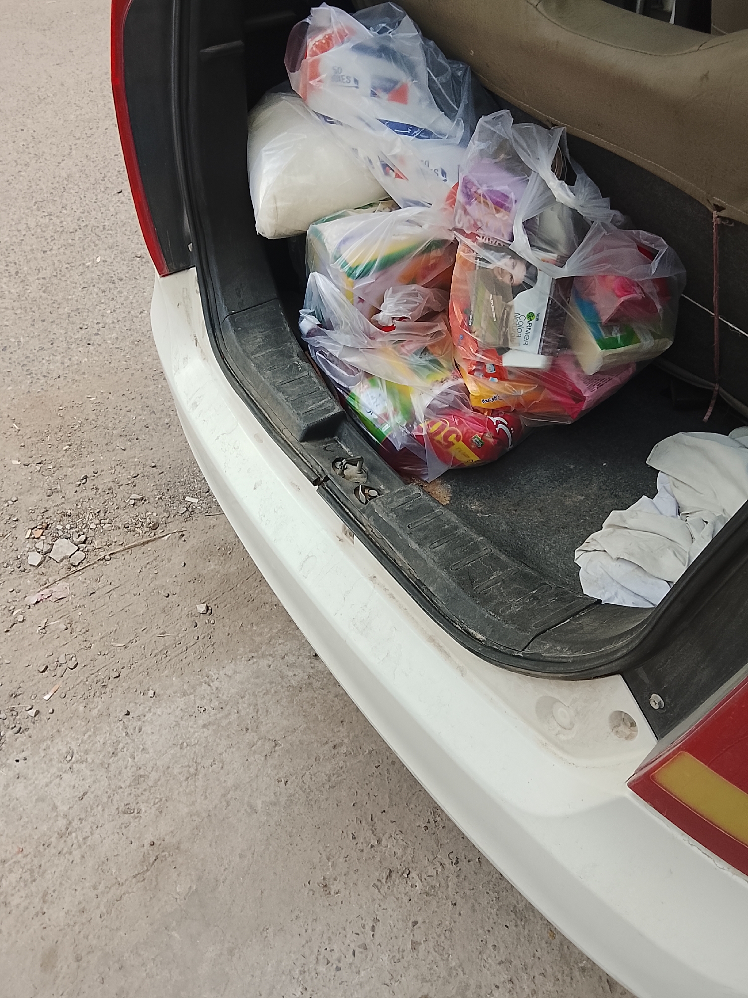 The open trunk of a white car is filled with grocery bags. The bags appear to contain a mix of food items and toiletries, with packaging visible including "Garnier Color Naturals" and other brands in what appears to be Urdu. The trunk lining is dark grey and slightly dirty. The vehicle is parked on a dusty surface, possibly a road or driveway, with debris scattered nearby. The location is possibly Gulgasht Colony, Pakistan, based on the packaging language and details of the request.