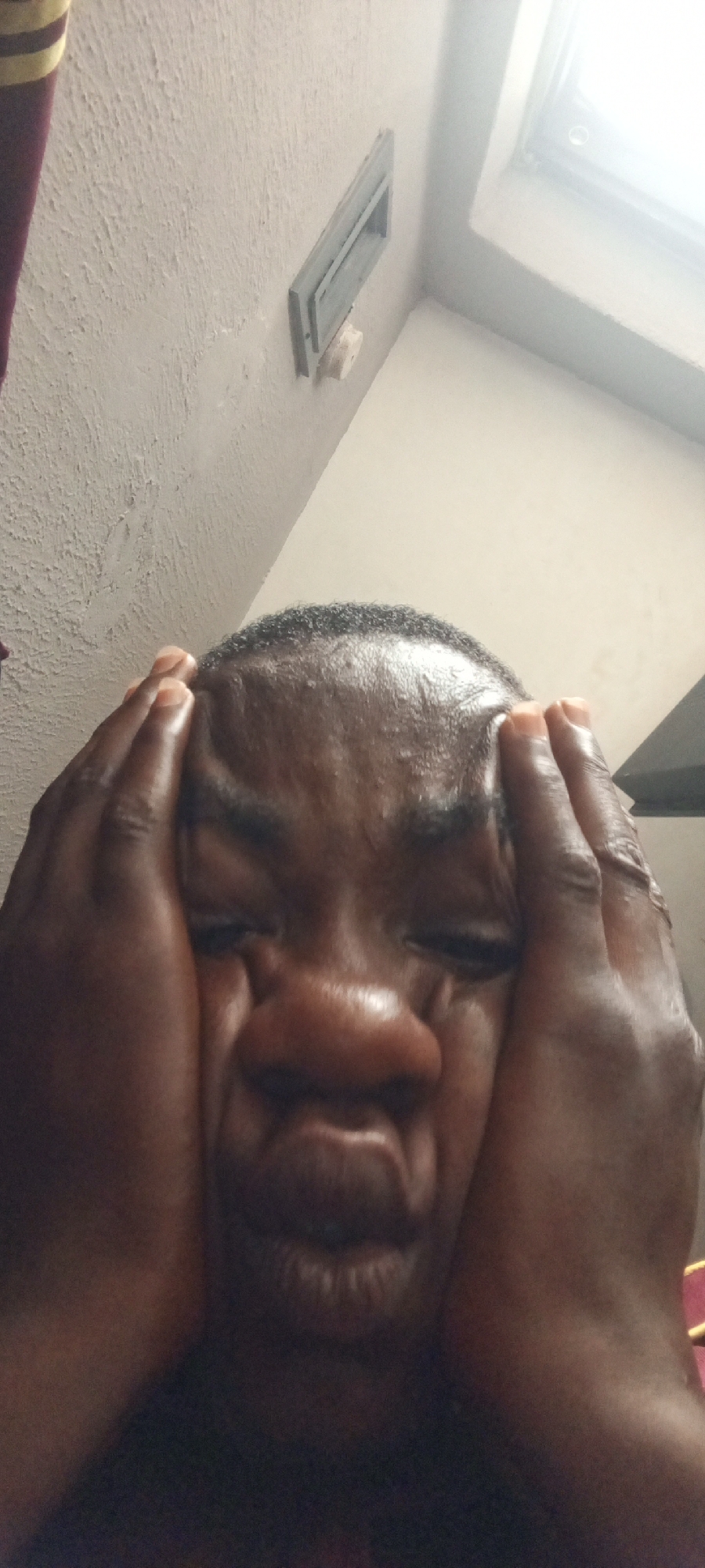 The image is a close-up, low-angle shot of a person's face with their hands pressed against their cheeks, distorting their features. The person appears to be an adult with dark skin and short, dark hair. Their eyes are closed or squinting, and their mouth is pursed. The background is a textured, off-white wall with a rectangular ventilation grate and a small white object mounted below it. A sliver of what appears to be a red and yellow striped fabric is visible in the upper left corner. The lighting suggests an indoor setting, possibly illuminated by natural light from a window or skylight, as indicated by the bright area in the upper right of the image. The scene appears to be within a room in a building located in Ilado, Nigeria.