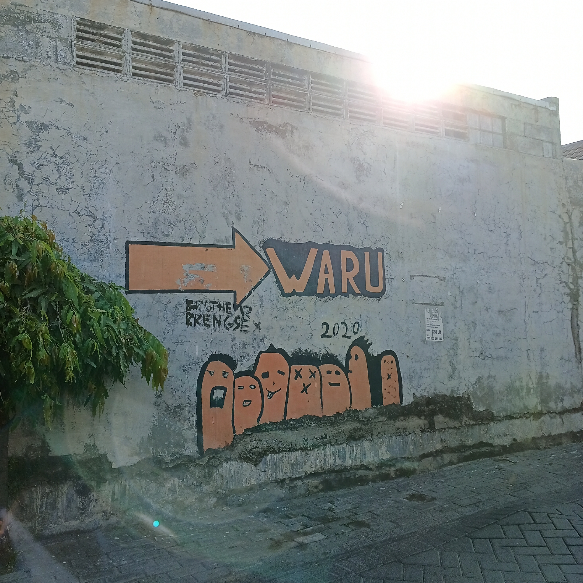 The image depicts a section of a weathered, light-colored concrete wall with graffiti. The graffiti features an orange arrow pointing to the right, with the word "WARU" painted in large, orange block letters outlined in black. Below the arrow, in smaller black lettering, are the words "BROTHER PRENGSE" and the year "2020." At the bottom of the wall, a line of stylized, orange figures with black facial features is painted. These figures appear to be anthropomorphic, with varying expressions, including one with crossed eyes and another with its tongue sticking out. To the left of the graffiti, a leafy green tree branches out, partially obscuring the wall. The wall itself shows signs of age, with cracking and discoloration. The ground in front of the wall is paved with rectangular bricks. Sunlight streams from the top right of the image, creating lens flare and casting shadows. A small, faded poster is affixed to the wall to the right of the "WARU" graffiti. The scene is located in the city of Wedoro, Indonesia.