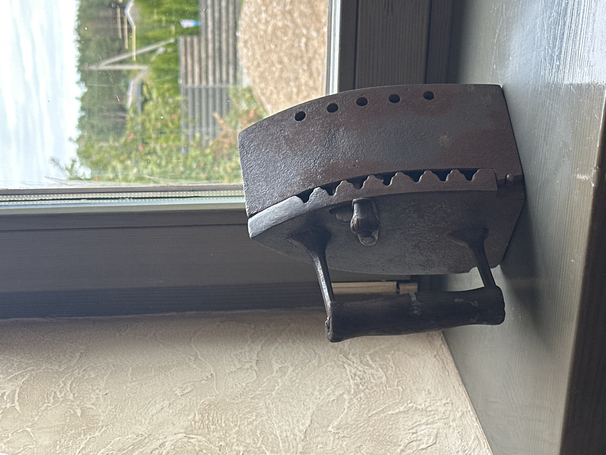 An old, rusty cast-iron flatiron sits on a gray windowsill in front of a window. The flatiron has a wooden handle and a serrated edge along one side. Through the window, a blurred view of trees, a fence, and a cloudy sky is visible. The wall to the left of the flatiron has a textured, off-white finish. The scene appears to be indoors, with the windowsill suggesting a domestic setting, possibly a house in the rural area of Нарацкі сельскі Савет, Belarus. There are no people or any discernible actions taking place.