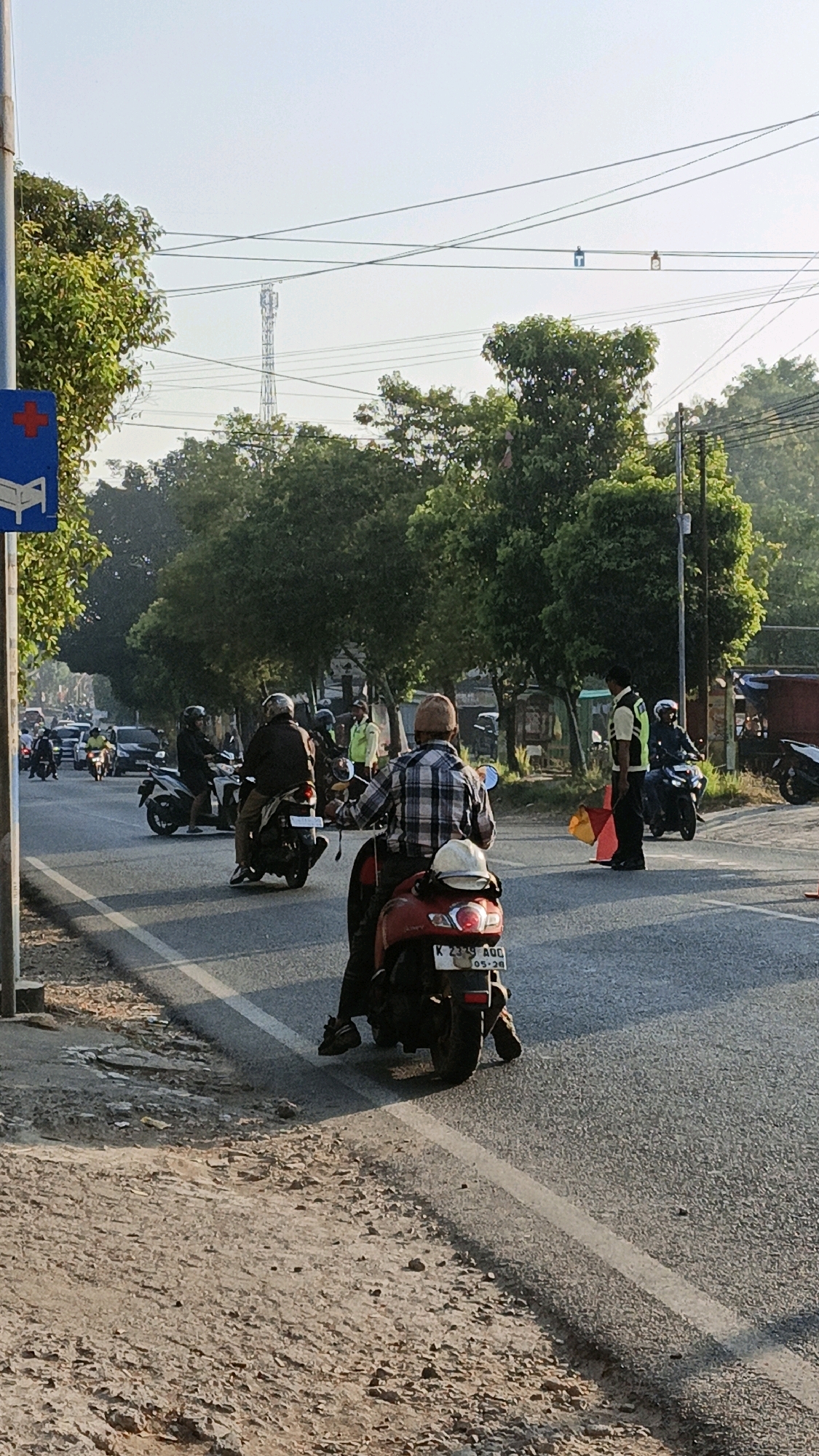 The image shows a street scene in Kuwasen, Indonesia, likely during the morning or late afternoon due to the quality of light. A road with a white line marking is visible, bordered by a dirt and gravel shoulder on the left and trees and vegetation on the right. Several people on motorcycles are on the road, navigating traffic. In the foreground, a person wearing a plaid shirt is riding a red scooter. Further down the road, a person in a reflective vest appears to be directing traffic, holding an orange and red traffic cone. Other motorcycles and some cars are present in the background, indicating moderate traffic. Overhead, power lines crisscross the sky, and a telecommunications tower is visible in the distance. A blue traffic sign with a red cross and a hospital bed symbol is mounted on a pole on the left side of the road. The overall impression is a typical urban street with active traffic and daily commuting.