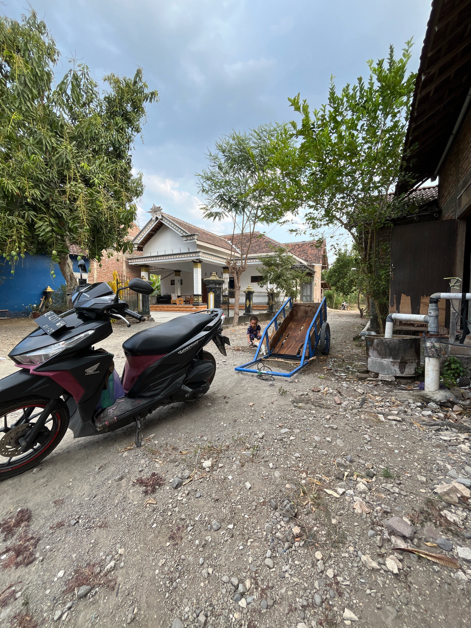 The image depicts an outdoor scene in the city of Kauman, Indonesia, likely a residential yard or common area. In the foreground on the left, a black and maroon Honda motorcycle is parked on a gravelly surface. To the right of the motorcycle is a wooden cart with blue metal framing, positioned on the ground. A young child, wearing a dark patterned shirt and shorts, is seated in the cart. Behind the cart and motorcycle, a white house with a distinctive ornate roofline is visible, flanked by trees. To the right of the house, a dark brown wooden structure with a corrugated roof is present, with plumbing pipes and a water barrel nearby. The sky is overcast with clouds.