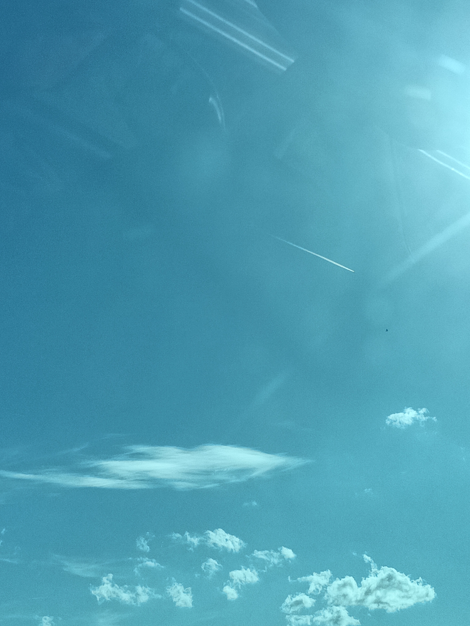 The image is a photograph of a bright blue sky with scattered clouds. A thin, white contrail from an airplane stretches diagonally across the upper middle portion of the frame. Towards the right side of the frame, a faint lens flare is visible. In the lower part of the image, several fluffy white clouds are dispersed. The overall scene suggests an aerial view or a clear day with an airplane passing overhead. The location is Divišov, Czechia.
