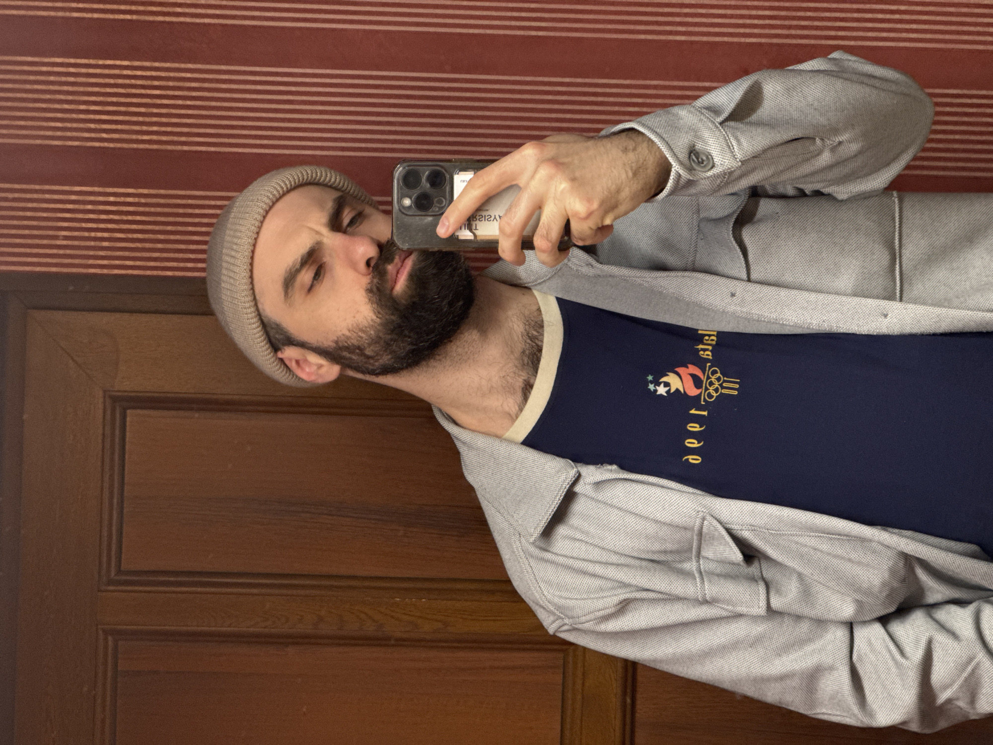 A man with a beard and a light brown beanie is taking a selfie with his phone. He is wearing a navy blue t-shirt with a graphic design and a light gray button-up shirt. He is standing in front of a brown door on the left and a red and white striped wall on the right. No evidence suggests that this image was taken in Yerevan, Armenia.