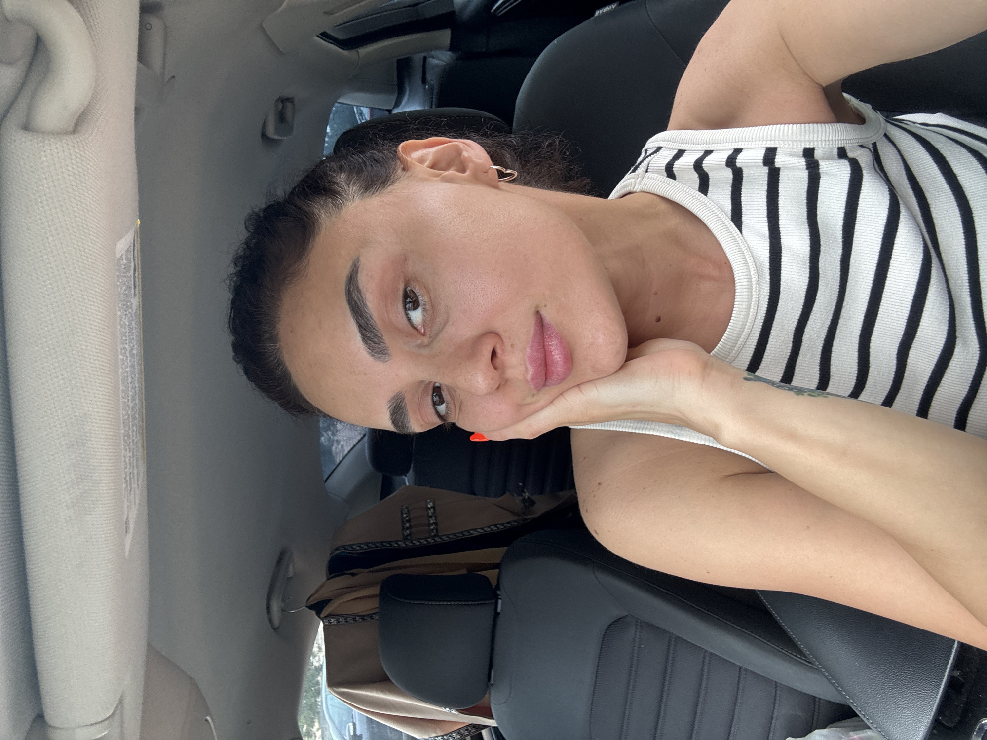 The image shows a woman sitting in the driver's seat of a car, possibly in Tbilisi, Georgia. She is wearing a black and white striped tank top and has her hand on her cheek. A brown jacket with ornate trim is hanging from the headrest of the passenger seat. The interior of the car is visible, including the dashboard and ceiling. The woman is looking at the camera.