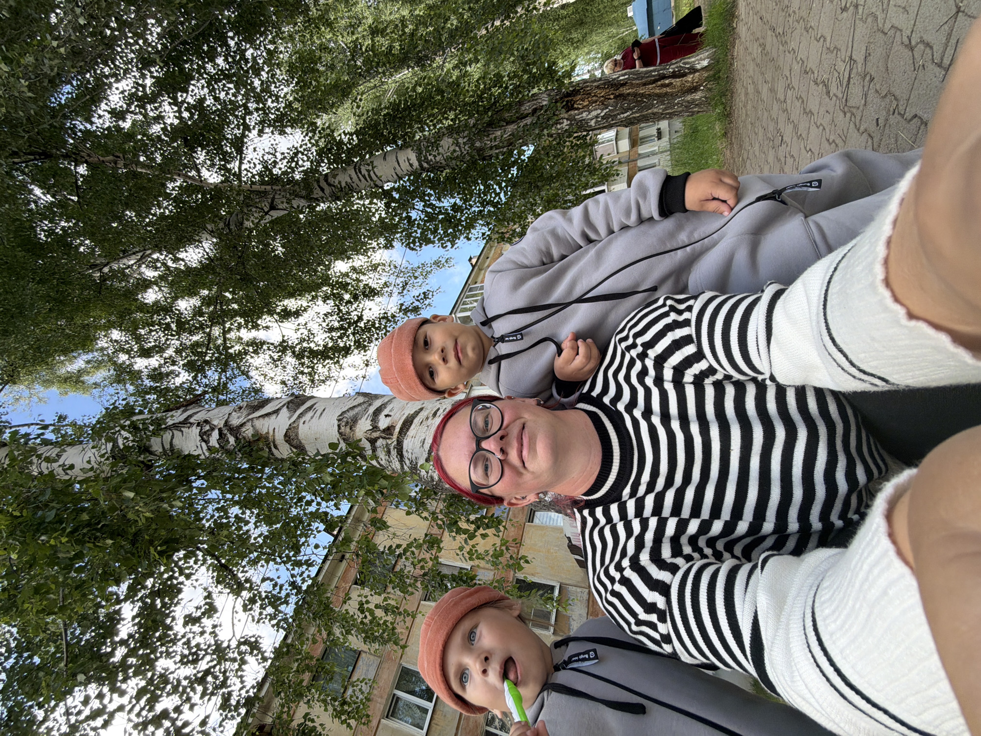 This is a close-up, low-angle selfie taken outdoors in Chaikovsky, Russia, likely on a sidewalk in front of a residential building. In the foreground, a woman with glasses and bright red hair is taking the photo, wearing a black and white striped sweater. To her left, a young boy in a grey onesie and an orange hat is visible, holding a green toothbrush to his mouth. To the woman's right, another young boy, also in a grey onesie and orange hat, is partially visible. Behind them, a birch tree with distinctive white bark and green leaves dominates the scene, with a multi-story residential building visible through the foliage. The sky is partly cloudy. In the background, on the right side of the image, another woman in a red top and dark pants is walking away from the camera. The ground is paved with light-colored interlocking bricks.