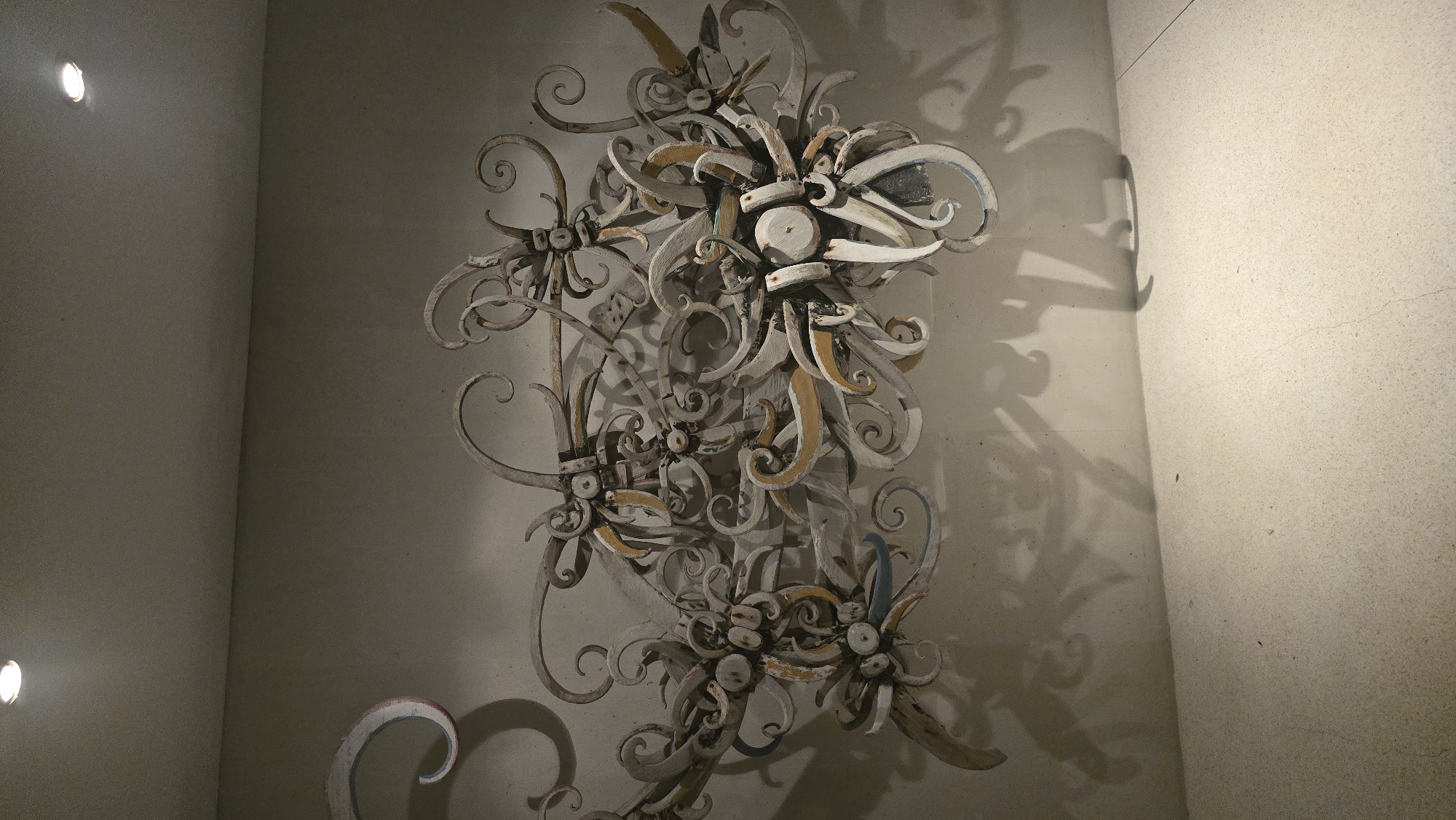 A low-angle, full-frame shot shows an ornamental ironwork or woodwork design mounted on a wall in a room. The piece consists of numerous swirling, repeating floral motifs and curved elements arranged vertically. The artwork has a weathered appearance with visible signs of wear on its painted surfaces. The colors of the artwork include white, beige, and touches of blue and gold. The wall is a pale beige color, and a shadow cast by the piece is visible to the right, suggesting a light source on the left. A spotlight illuminates the wall at the top left. The bottom left shows a partial curved element painted with pink and white. The location is given as Kuta Selatan, Indonesia.