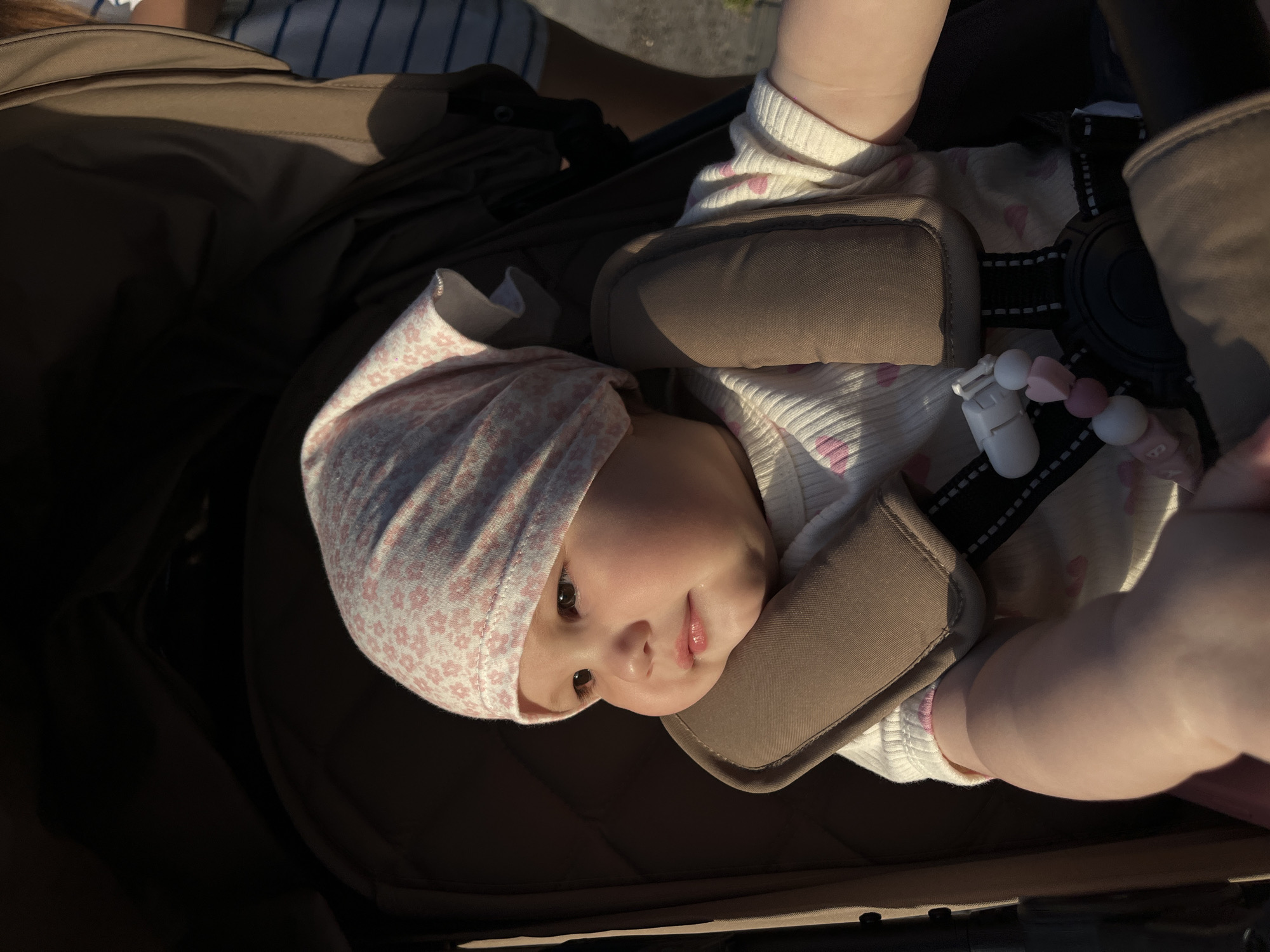 A baby wearing a pink and white floral bonnet and a white shirt with pink hearts lies in a brown stroller. The stroller has brown padded straps and a black buckle. A pacifier clip with white and pink beads is attached to the stroller strap. The baby is looking directly at the camera. A person wearing a blue and white striped shirt is partially visible in the top left corner. The scene appears to be outdoors. The location is unspecified and unrelated to Novopokrovskaya, Russia.