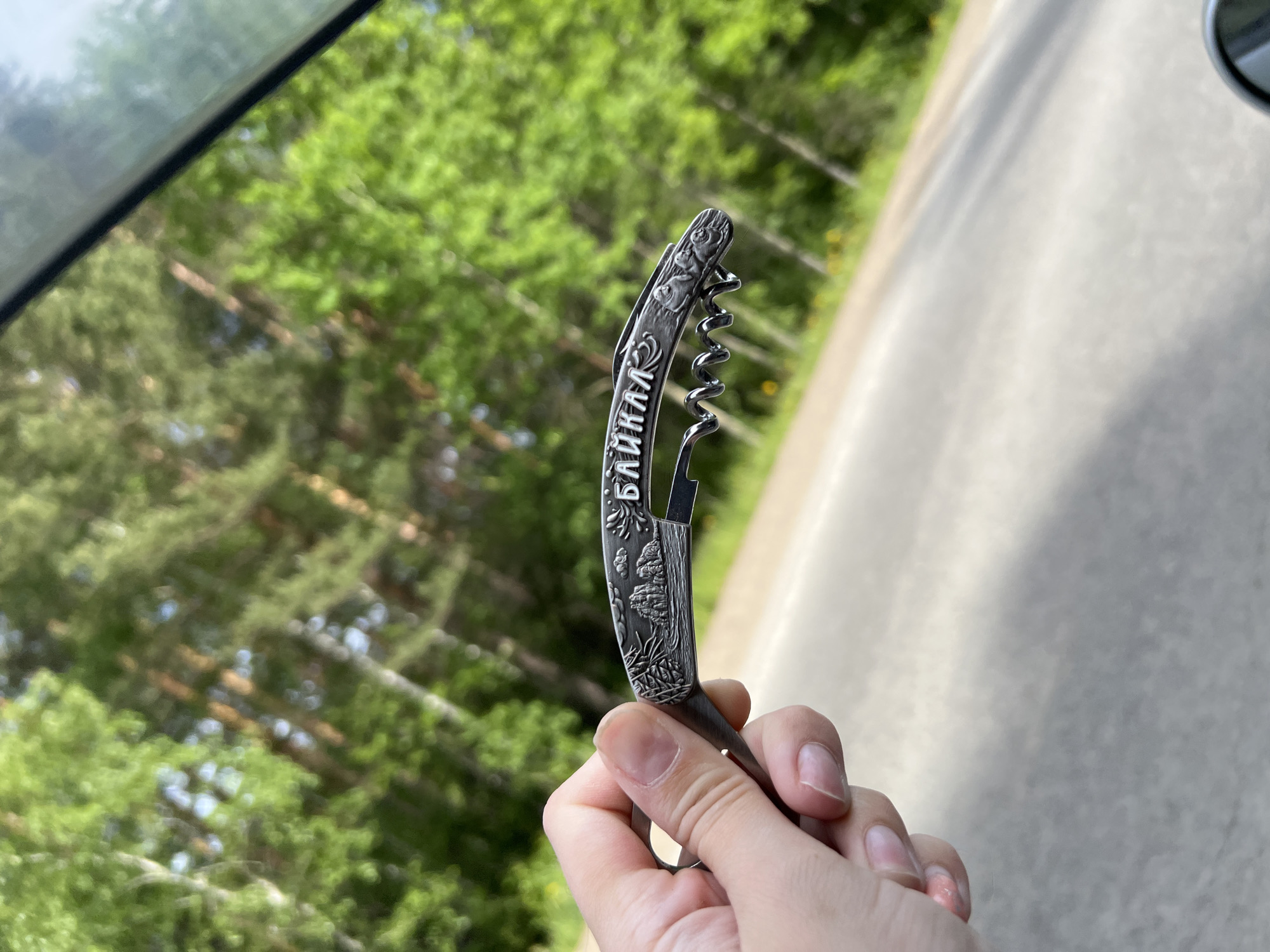 A person's hand holds a metallic corkscrew with intricate carvings. The corkscrew displays a landscape scene on its handle, featuring mountains, trees, and clouds. Text in Cyrillic letters, appearing to read "БАЙКАЛ," is inscribed on the handle. The corkscrew is positioned against a backdrop of lush green trees, suggesting an outdoor setting. The image was captured in the vicinity of Pivovarikha, Russia. The background shows a paved road and a blurred view of trees, possibly indicating a roadside or a forest edge.