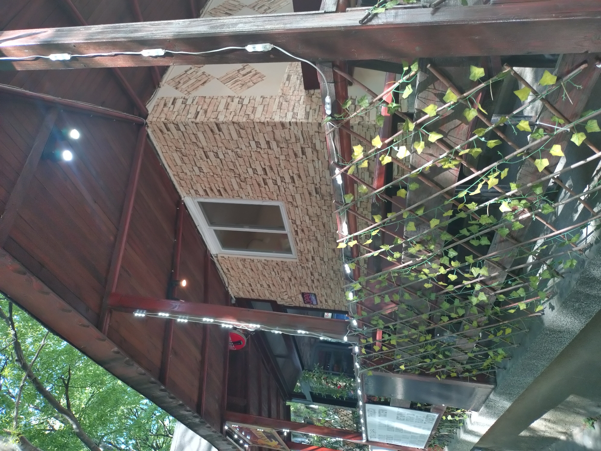Here's a description of the scene:
The image shows an outdoor structure, possibly a restaurant or cafe, located in Stara Zagora, Bulgaria. It is an upward shot showing the underside of the structure's roof and ceiling. The roof is wooden, with string lights attached. A section of the ceiling is covered in brick-patterned material and includes a window. A decorative lattice with artificial vines covers another portion of the ceiling. A menu board and other signage are visible below, suggesting this is a commercial establishment. The tree branches and sky can be seen in the bottom left corner.