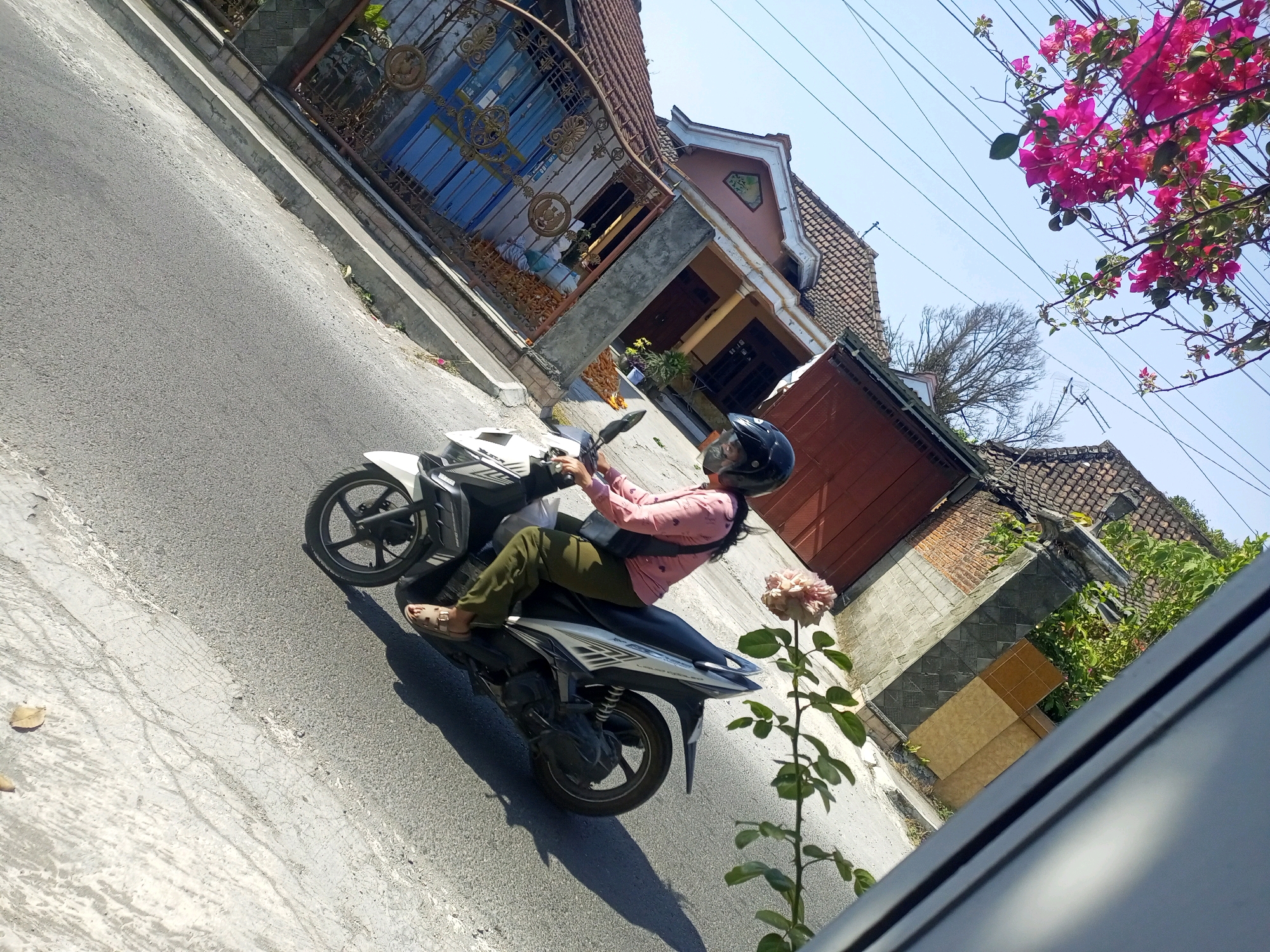 The image shows a person riding a white motorcycle on a street in Ngasem, Indonesia. The rider is wearing a pink shirt, green pants, and a black helmet. A pink flower is visible in the foreground on the right side of the image. Behind the motorcycle, there are houses with terracotta tiled roofs and a blue gate. Overhead, there are power lines and a bright blue sky. The street appears to be paved with asphalt and has a concrete curb.
