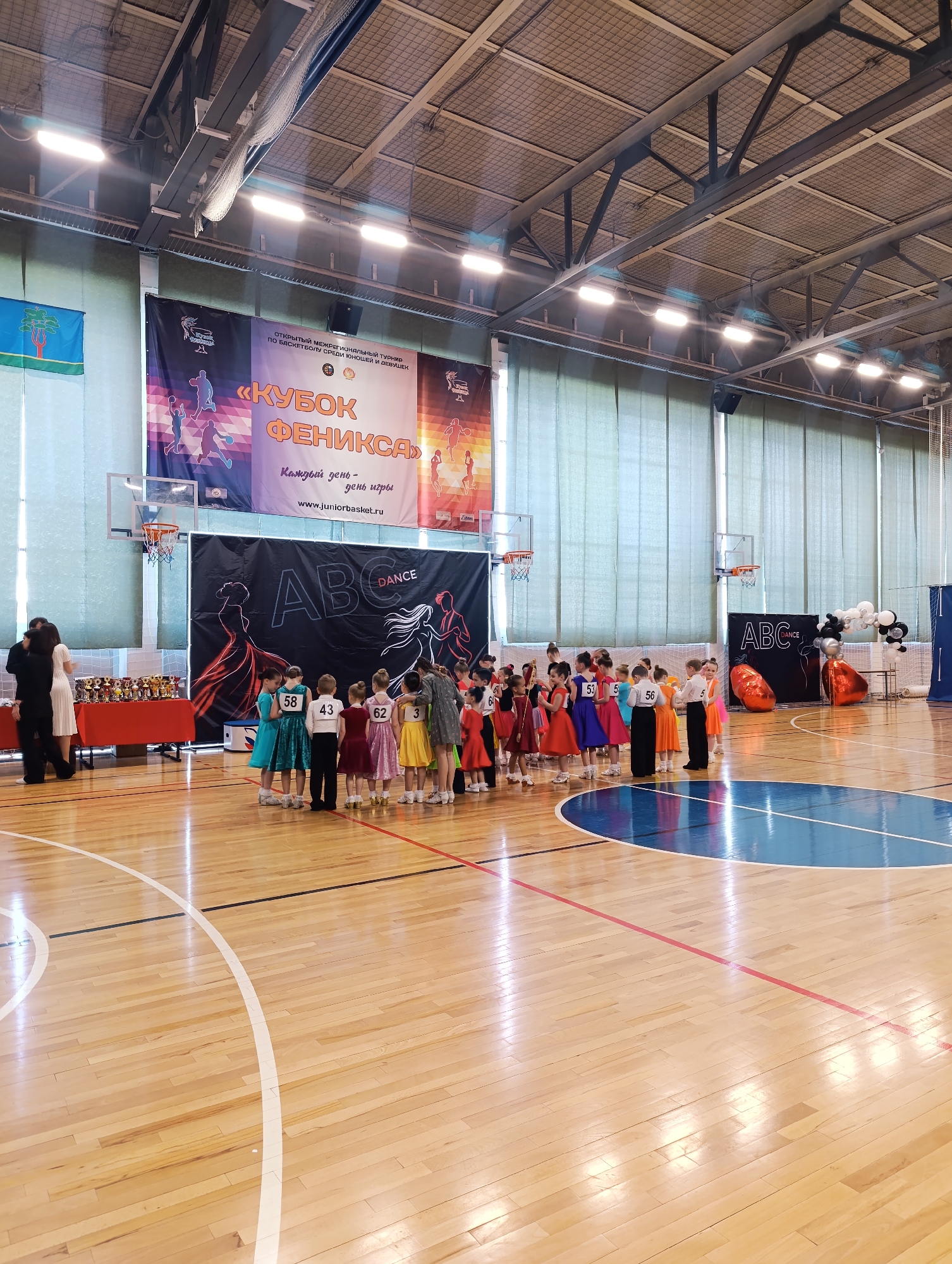 The image depicts an indoor sports arena, possibly in the city of Unknown, Unknown. A group of children, presumably dancers, are gathered in the center of the wooden basketball court. They are wearing formal attire, primarily dresses and suits, and numbered bibs. A woman in a grey dress is interacting with the children.
Behind the group is a banner with "ABC DANCE" printed on it, flanked by large orange balloons. Banners with the text "КУБОК ФЕНИКСА" are hung on the background wall. To the left, there is a table laden with numerous trophies and two people are standing near the trophies. The environment is an indoor arena with visible basketball hoops, and the architecture includes ceiling supports and lighting fixtures.