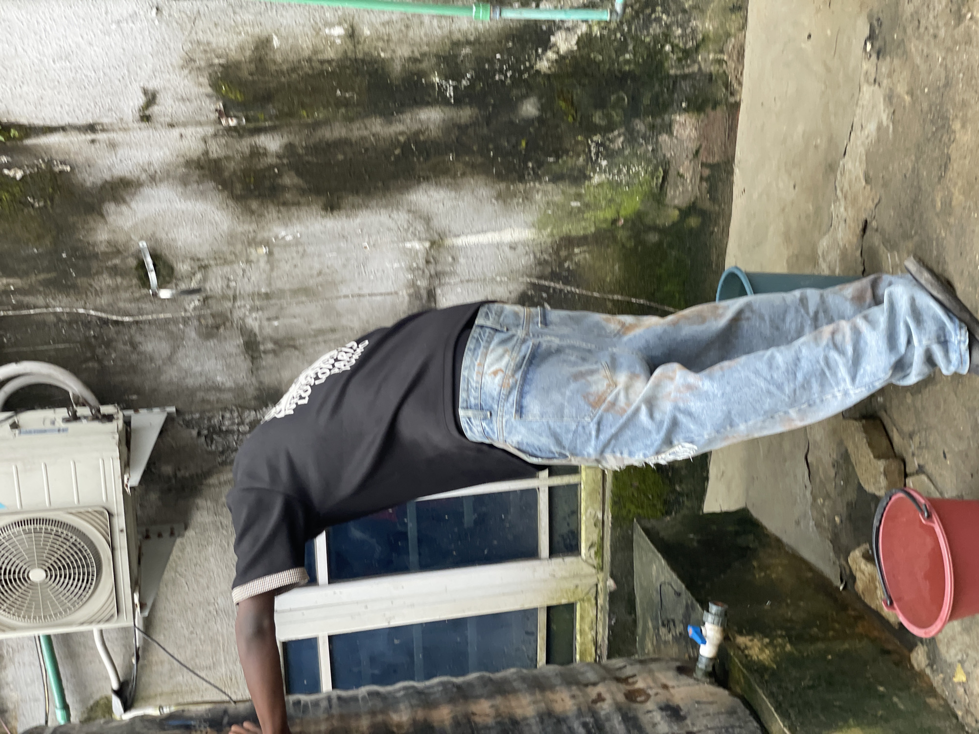 A person wearing a black t-shirt and distressed denim jeans leans against a large, dark cylindrical object, possibly a water tank, with their left arm resting on it. The person's face is not visible as their head is turned away from the camera and bent downwards. They appear to be wearing dark-colored shoes.
The background consists of a textured, grimy concrete wall, stained with dark patches and moss. An air conditioning unit is mounted high on the wall above the person. To the right, a bright green pipe runs vertically along the wall.
In the foreground, a red bucket sits on the ground to the left, near the base of the cylindrical object. A blue faucet is attached to the side of the cylindrical object, and it appears to be dripping or has recently been used, as there is a puddle of water on the ground beneath it. To the right of the person, a teal-colored bucket is positioned on the ground.
The scene appears to be an outdoor area, possibly a residential or commercial space, with elements suggesting a functional water supply system. The environment seems weathered and somewhat neglected, with visible signs of moisture and algae growth on the surfaces. The lighting suggests it might be daytime, but the overall atmosphere is muted. The location is specified as Ado, Nigeria.