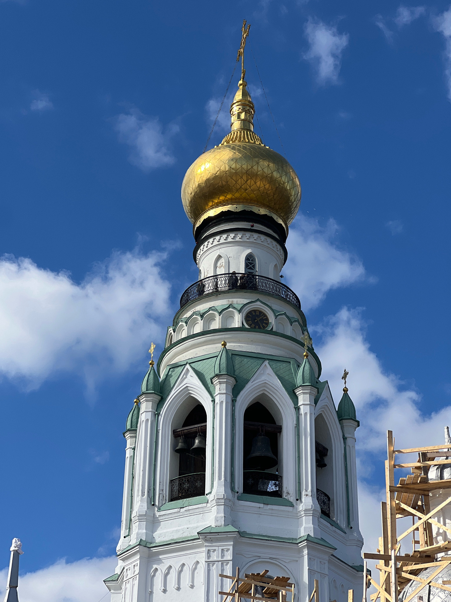 This is a close-up, low-angle, eye-level, outdoor, daylight, full-color, medium shot photograph of a white bell tower with green roofs and a golden dome. The tower has multiple levels with arched openings where bells are visible. A clock face is also visible on one of the levels. The tower is adorned with crosses. A wooden scaffolding is present on the right side of the frame. The sky is blue with scattered white clouds. There are no people or animals in the frame.