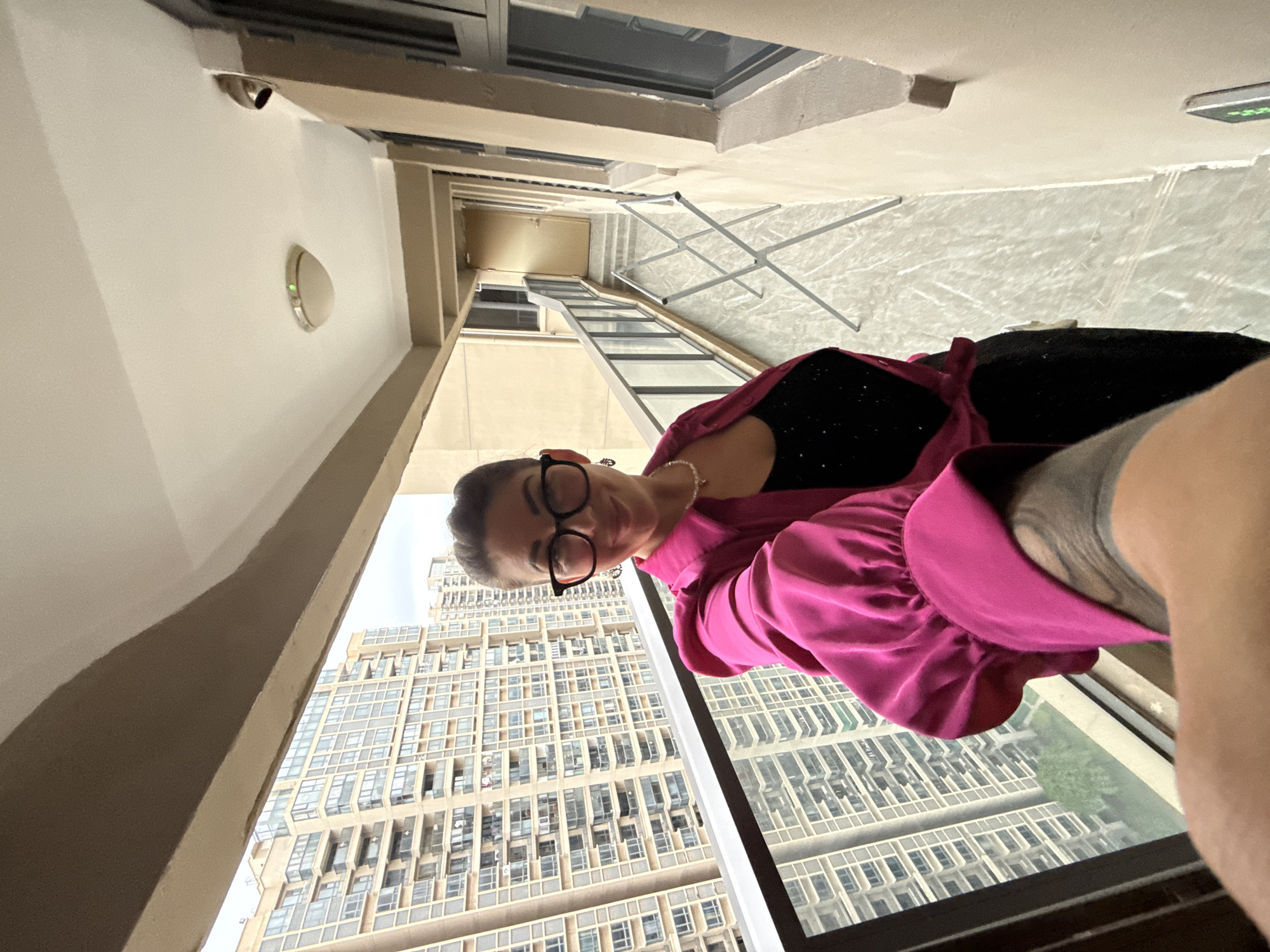 A woman is taking a selfie on a balcony, with a high-rise apartment building in the background, likely in Huizhou, China. The woman is wearing a pink, ruffled blouse and a black, sequined top. She has dark hair pulled back and is wearing glasses and earrings. The balcony has a glass railing and the floor appears to be tiled. In the background, beyond the railing, are multiple tall apartment buildings. The balcony extends to a doorway at the far end, with a clothes drying rack visible in the middle. The lighting suggests it is daytime, with overcast skies.