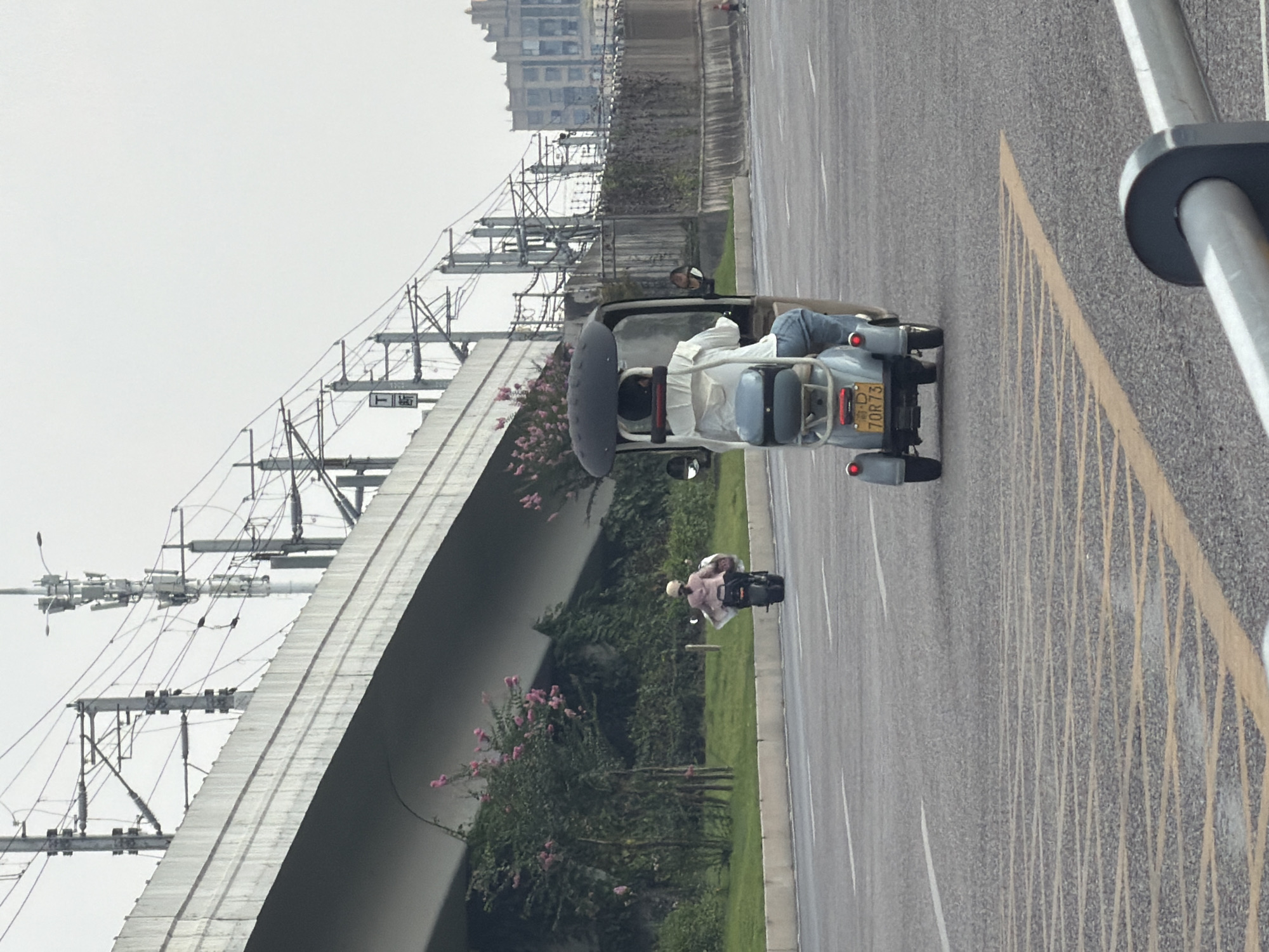 A three-wheeled scooter with a canopy is being operated by a person wearing a white shirt and blue jeans. The scooter is traveling on a paved road, as indicated by the yellow lines and white lane markers. In the background, there is an elevated concrete structure with overhead power lines and communication towers. Vegetation, including trees with pink flowers, lines the side of the road. Further in the distance, a multi-story building with a distinctive facade is visible. Another person on a motorcycle is further ahead on the road. The scene appears to be in an urban or semi-urban environment, possibly in Jiangjin, China, given the presence of the elevated transport infrastructure and the style of the vehicles. The sky is overcast.