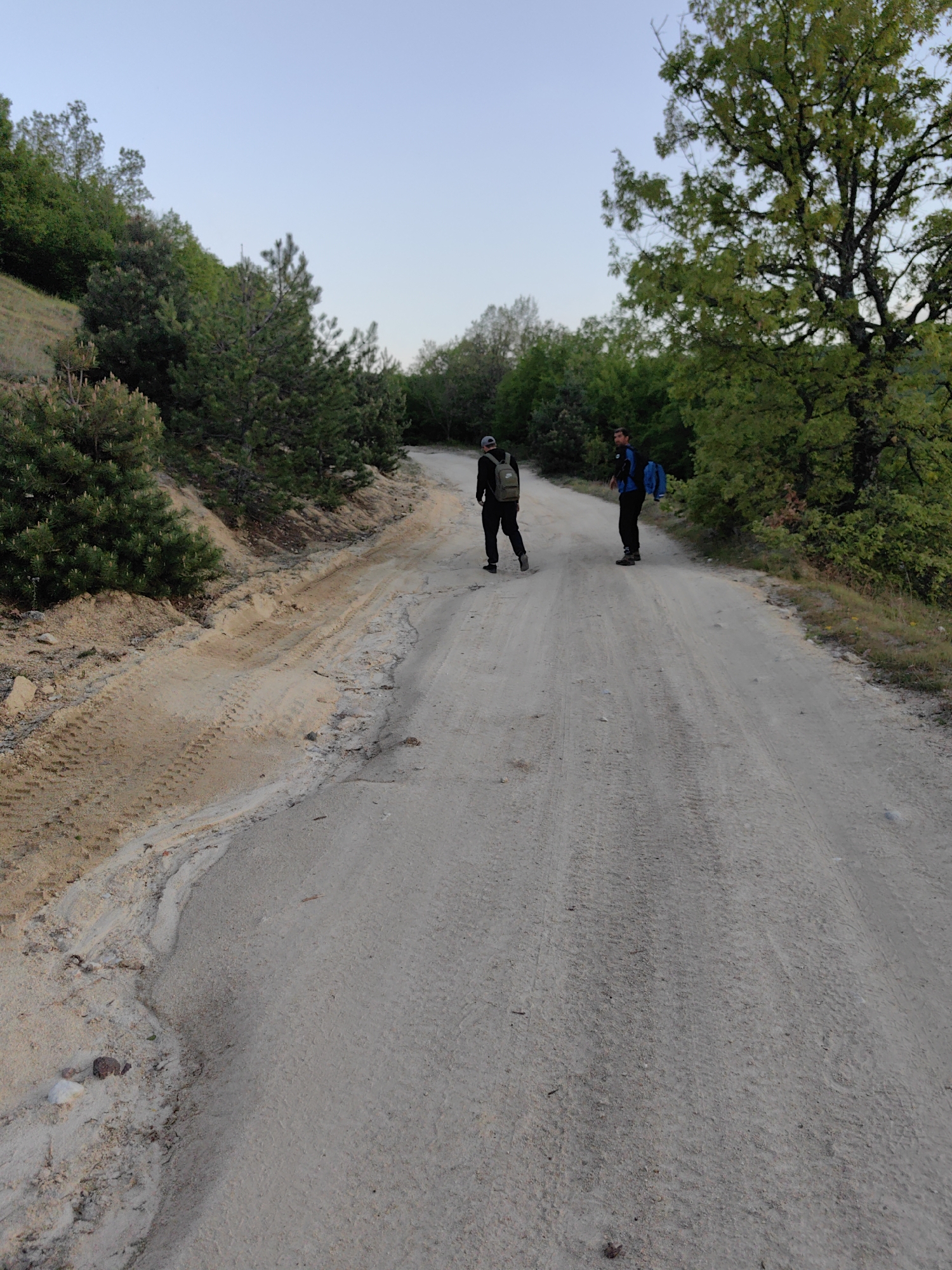 The image depicts two individuals walking on a dirt road in a rural environment. The road appears to be unpaved and is flanked by trees and vegetation. The sky is visible in the background. The individuals are wearing casual clothing and backpacks, suggesting they are hiking or walking. The scene is observed during daylight. The location is Hristo Danovo, Bulgaria.