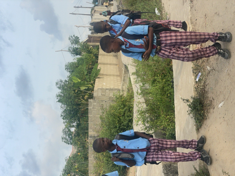 The image depicts an outdoor scene in Mosafejaw, Nigeria, featuring three young male individuals, identified as students by their uniforms, standing on a path under an overcast sky.
The uniforms consist of light blue short-sleeved shirts, maroon and white striped ties, red and white plaid trousers, and dark closed-toe shoes. Each student wears a black backpack. The individual on the far left stands facing right, his left hand in his trouser pocket, looking towards the central figure. The central individual stands facing left, arms crossed over his chest, wearing a gold-buckled belt, and looking towards the left individual. The third student is positioned behind and to the right of the central figure, partially obscured, also facing left. Several indistinct figures are visible in the far background on a dirt path to the right.
The immediate ground transitions from a concrete slab on the left to a dirt path on the right. Green bushes line both sides of the path. A grey concrete block wall extends across the midground, behind which abundant green vegetation, including tall, slender trees and broad-leafed plants consistent with banana or plantain trees, is visible. A dark metal gate is discernible behind the wall on the right. Utility poles with overhead wires are present in the far right background. White debris, possibly paper or plastic, lies on the dirt path in the foreground. The sky is largely covered by grey and white clouds.