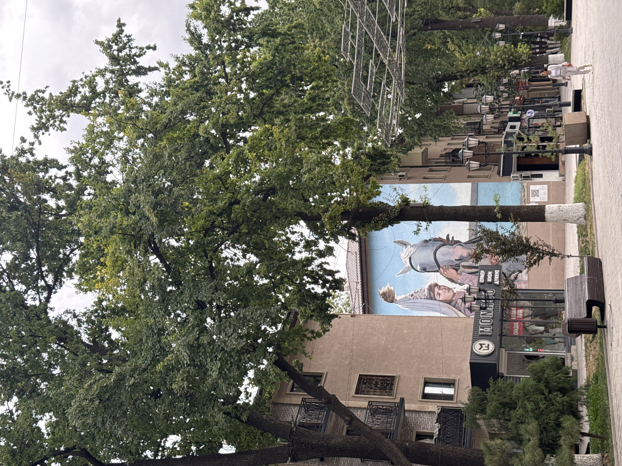 The image depicts a street scene in Shymkent, Kazakhstan, dominated by large, leafy trees. In the foreground, a paved walkway runs alongside a grass verge. A dark brown park bench is situated on the walkway, adjacent to a trash receptacle. To the left of the bench is a shop with a black awning and the name "MONACO" in white lettering. The shop window displays clothing and signs indicating sales. A mural of a person and a horse is painted on the building behind the shop. A woman in a light-colored suit is walking away from the viewer on the walkway to the right. The background features buildings and numerous lampposts lining the street, suggesting an urban environment. The sky is overcast.