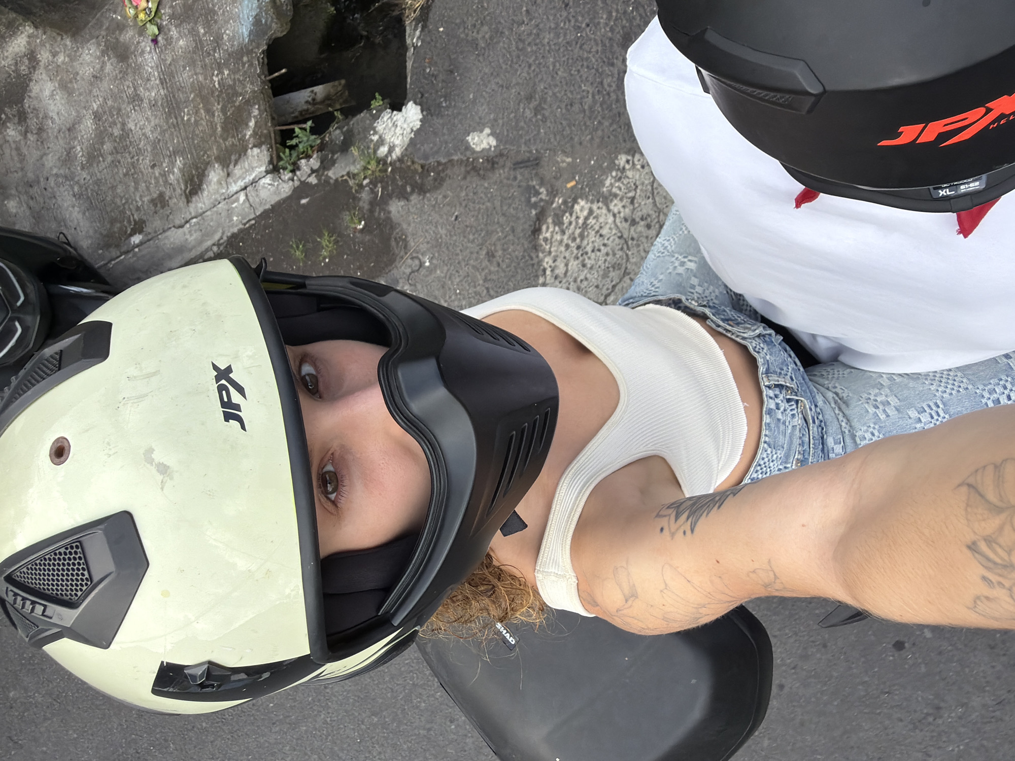 A person wearing a cream-colored JPX helmet and a white tank top is seated on a dark-colored scooter, looking directly at the camera. The person has brown eyes and curly hair. They are wearing light blue jeans with a patterned texture. Their left arm is visible, showing tattoos of floral designs. The scooter is parked on a paved surface, likely a street, with a gutter to the right containing some greenery and debris, consistent with locations in North Kuta, Indonesia. Another helmet, black with red accents and "JPX" logo, is partially visible in the lower right corner. The overall lighting suggests it is daytime.