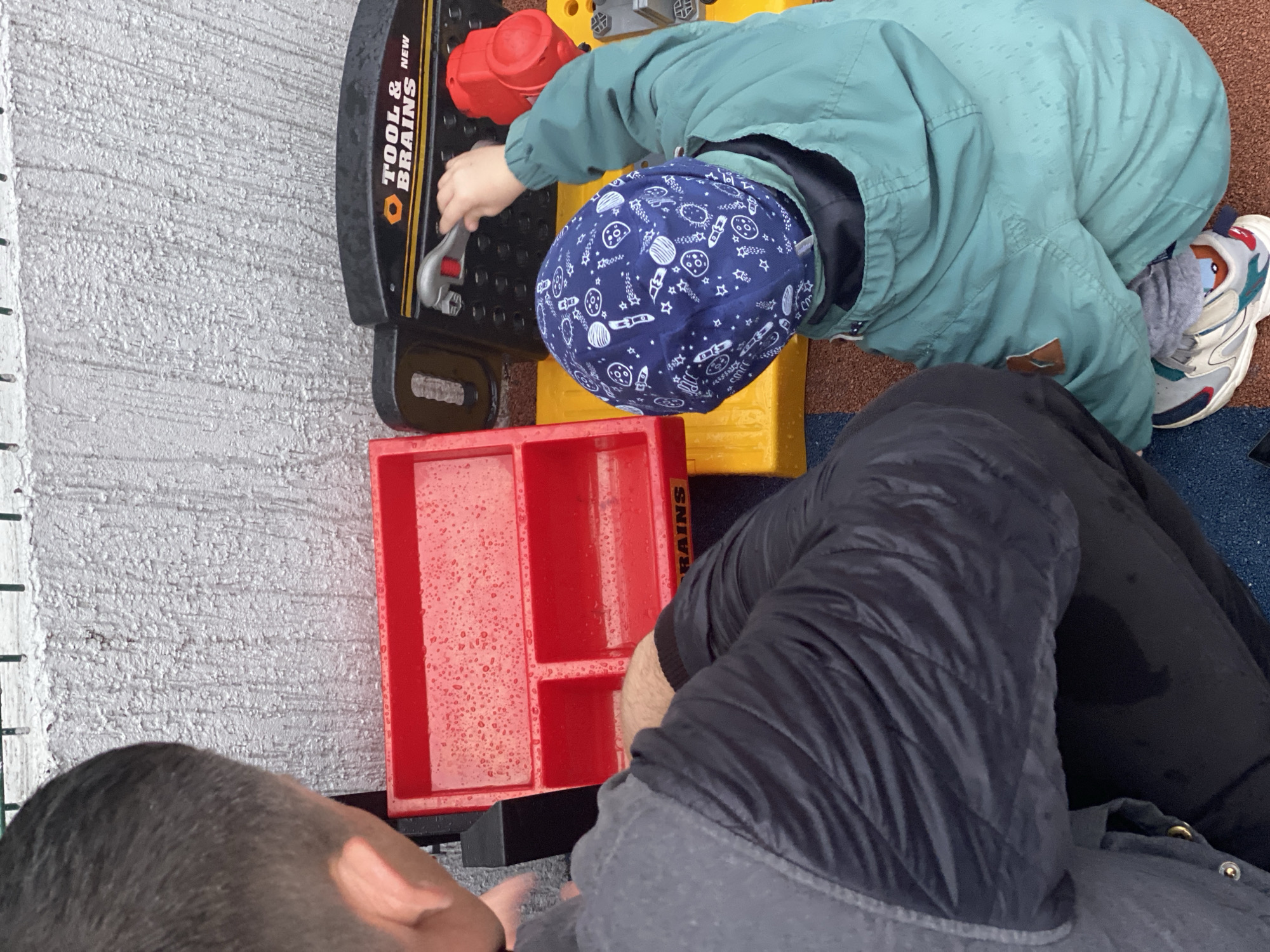 A child wearing a blue hat patterned with white celestial objects (planets, stars, rockets) and a light green jacket is playing with a "TOOL & BRAINS" branded toy workbench, which includes a red toolbox and a red knob. The child is using a toy wrench. The scene also includes a man wearing black pants and a gray jacket. The location appears to be outside, possibly a playground or backyard. The location in the city of Sochi, Russia cannot be determined from the image.