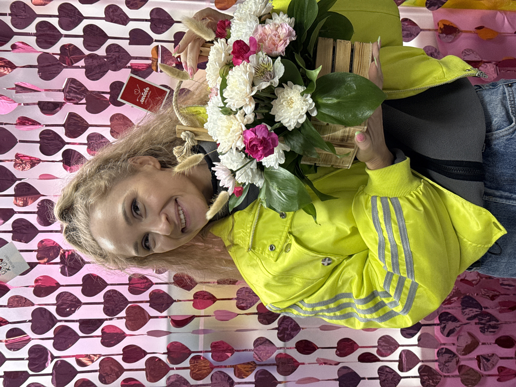 A smiling Caucasian woman with blonde hair holds a wooden basket of white and pink flowers against a backdrop of pink heart-shaped decorations. She is wearing a bright yellow jacket with gray stripes on the sleeve, a gray top, and blue jeans. A small red card with Russian text on it is visible near the flowers. The photo may have been taken indoors, possibly in Perm, Russia.