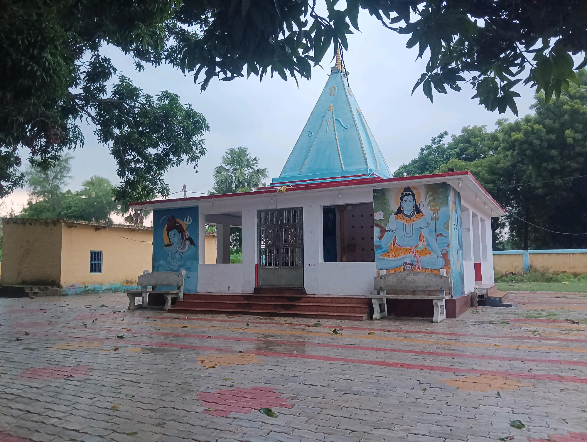 This is a full, eye-level shot of a small Hindu temple in Tilkar, India, on an overcast day. The temple is painted white with a light blue, pyramid-shaped spire topped with a gold finial. The spire is adorned with triangular symbols. The sides of the temple building are decorated with murals of deities. To the left, a mural depicts a blue-skinned figure with dark hair, holding a trident. To the right, a mural shows a similar figure, also with dark hair, seated and holding objects in its hands. A metal gate covers the entrance to the temple, and red-brown steps lead up to it. Two concrete benches with backrests are placed in front of the temple, one to the left and one to the right of the entrance. The ground in front of the temple is paved with interlocking bricks in a pattern of red, yellow, and gray. Fallen leaves are scattered across the paving stones. Behind the temple, a yellow building with a blue-barred window is visible. Trees with green foliage surround the temple complex. A low, yellow wall with a blue top runs along the back of the property. The sky is a dull, cloudy gray.
