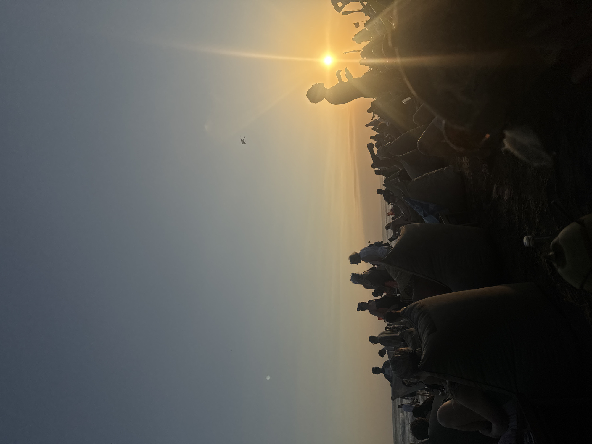 This image depicts a sunset gathering on a beach in North Kuta, Indonesia. The setting sun, positioned on the right side of the horizon, emits a strong bright yellow and orange light, creating significant lens flare and backlighting, which silhouettes most subjects. The sky transitions from a pale blue at the top to warm orange and yellow hues near the horizon.
Numerous individuals are present across the dark sandy beach. Many people are seated on large, dark-colored beanbags, particularly visible in the foreground and midground. One prominent person stands silhouetted against the intense sunlight on the right, appearing to gaze towards the horizon. Other individuals are standing or moving along the beachfront, with some visible near the water's edge on the far left, where gentle ocean waves are breaking. In the upper mid-sky, a dark, geometric kite is visible, flying. In the extreme lower right foreground, a partially visible coconut and a small plastic bottle are present on the sand. The overall scene suggests a relaxed communal event focused on viewing the sunset.