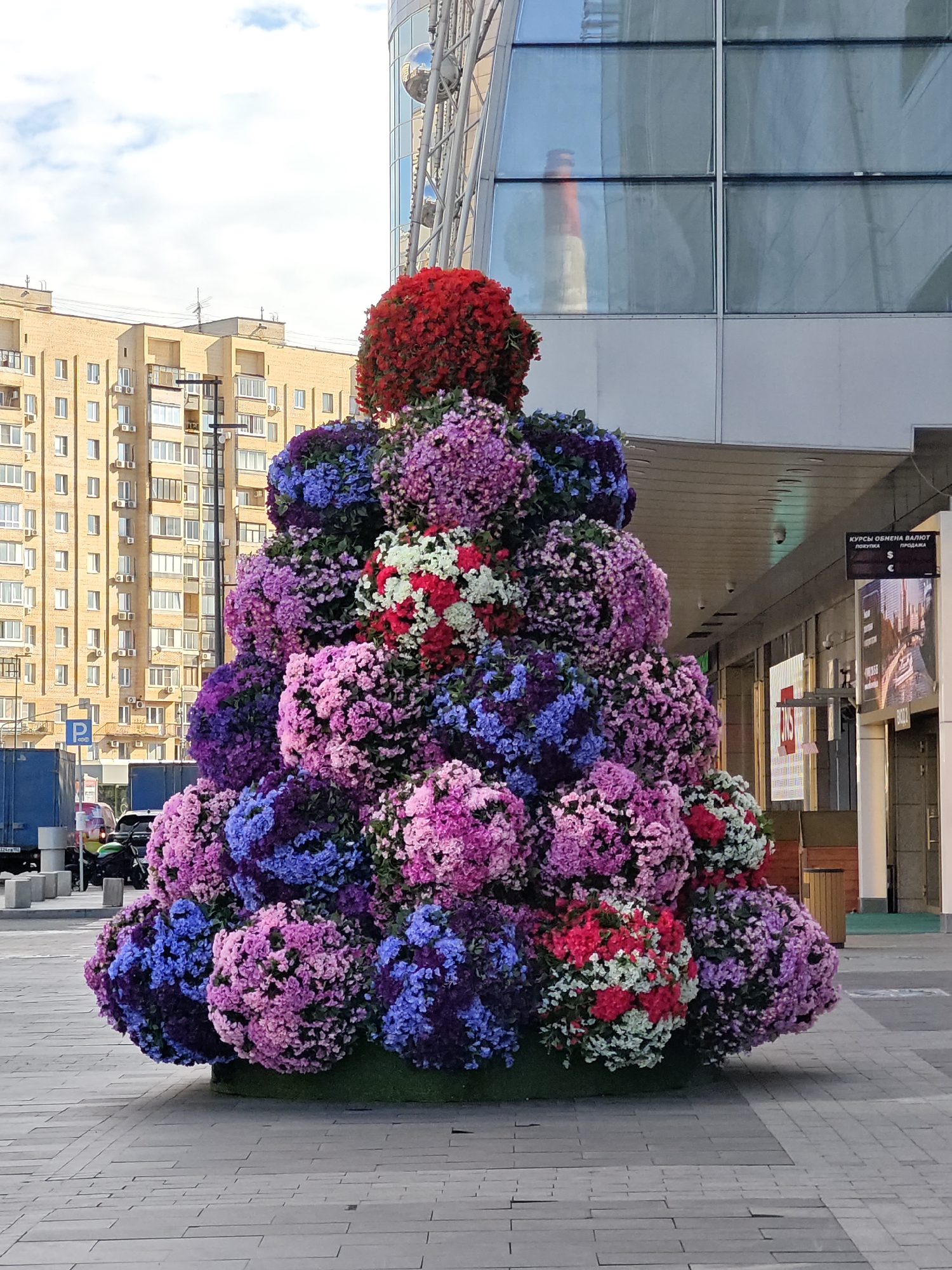 An outdoor, full-shot, eye-level, medium perspective photograph captures a large, pyramid-shaped floral arrangement situated on a paved walkway in front of a modern building. The arrangement is composed of numerous spherical clusters of flowers in various shades of purple, pink, blue, and red, with a dominant red cluster at the apex. Behind the floral display, a multi-story residential building with a light-colored facade and numerous windows is visible, along with a portion of a contemporary glass-fronted commercial building. To the left of the floral arrangement, a blue truck and a parked motorcycle are present. A blue "P" sign for parking is also visible. The scene appears to be in a public urban space in Moscow, Russia, likely a commercial or residential area. The sky is overcast with scattered clouds. No individuals are clearly depicted in the image.