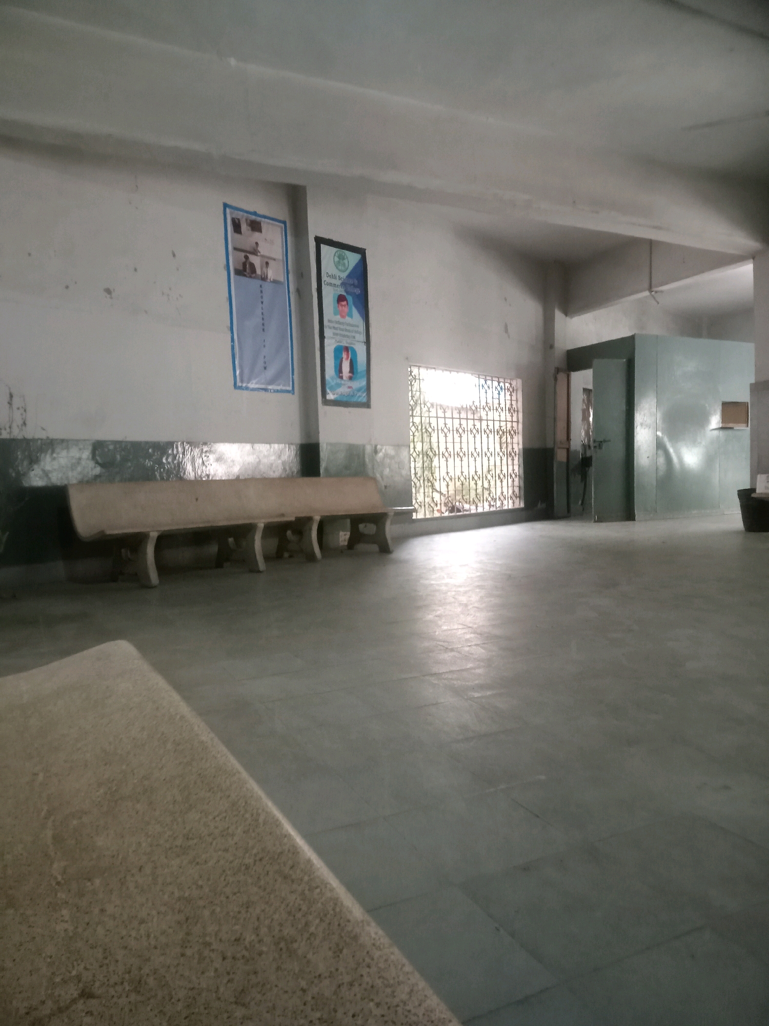 The image depicts an interior hallway or waiting area within a building. The walls are painted a light color and appear somewhat worn, with visible marks and a dark green wainscoting strip running horizontally. Two framed posters are affixed to the wall on the left. One poster is predominantly blue with text and images. The other, to its right, is a vertical poster with a green border, featuring a portrait of a person at the top and more text below.
A long, light-colored concrete bench with several supporting legs is positioned against the wall. In the background, a window with a decorative metal grate allows light to enter the space. To the right of the window, there is a large, dark green structure that appears to be a partition or a small room, with a door visible. Another smaller, light brown window or opening is set into this structure. The floor is tiled with large, square, dark gray tiles, which are reflective in the lighting. In the foreground, a portion of another light-colored bench or seating area is visible, angled towards the right side of the frame. The overall impression is of a utilitarian and somewhat aged public space. The specific location within Karachi Division, Pakistan, cannot be determined from the visual information alone.