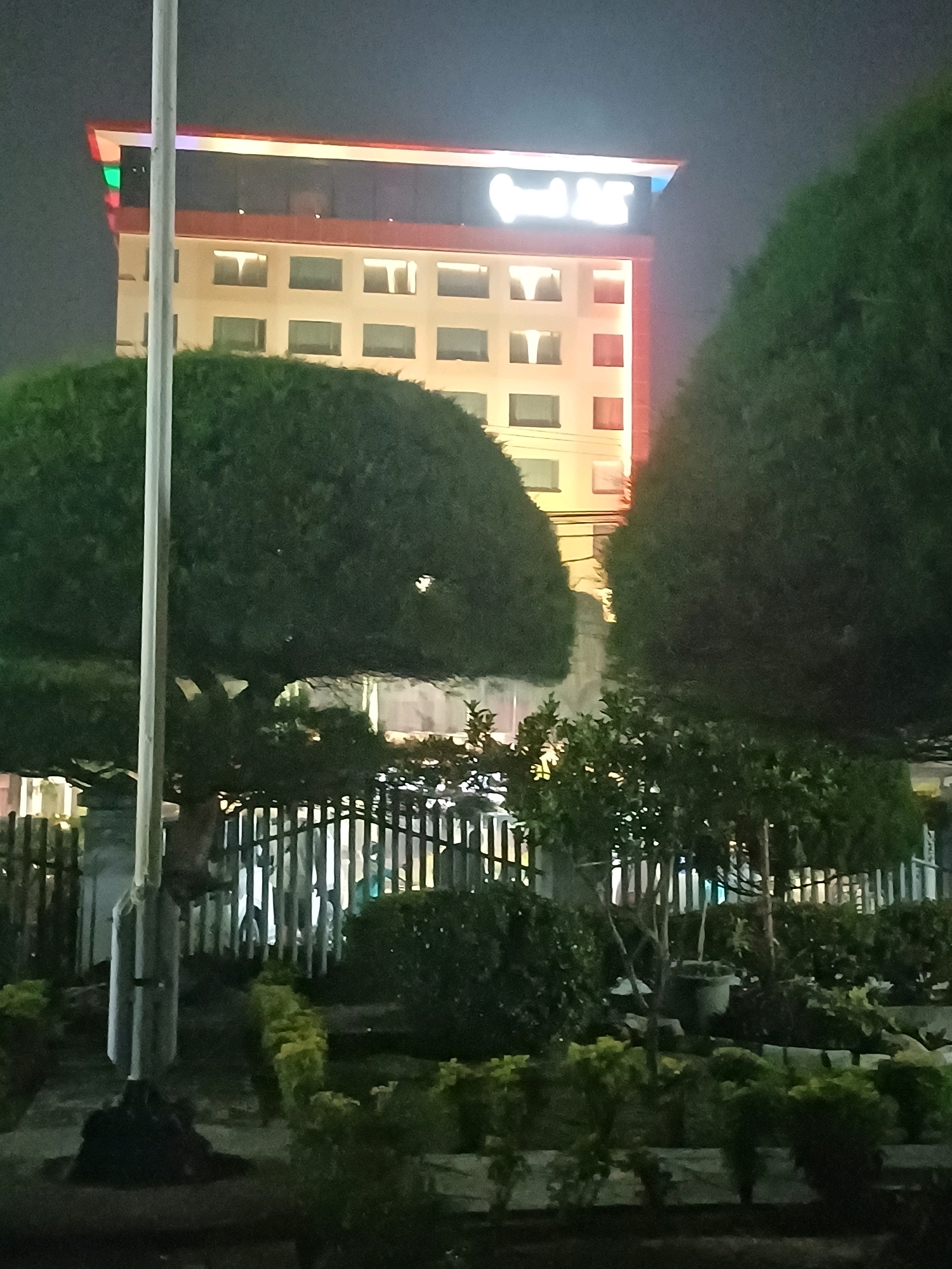 The image shows a building at night in Dumai City, Indonesia. The building is tall and illuminated with colorful lights on top. The facade is light-colored with multiple windows and some orange accents. In front of the building, there are large, rounded trees and a dark fence. The foreground includes a lamppost and dense foliage. The overall scene suggests an urban environment at night, possibly a hotel or commercial establishment.
